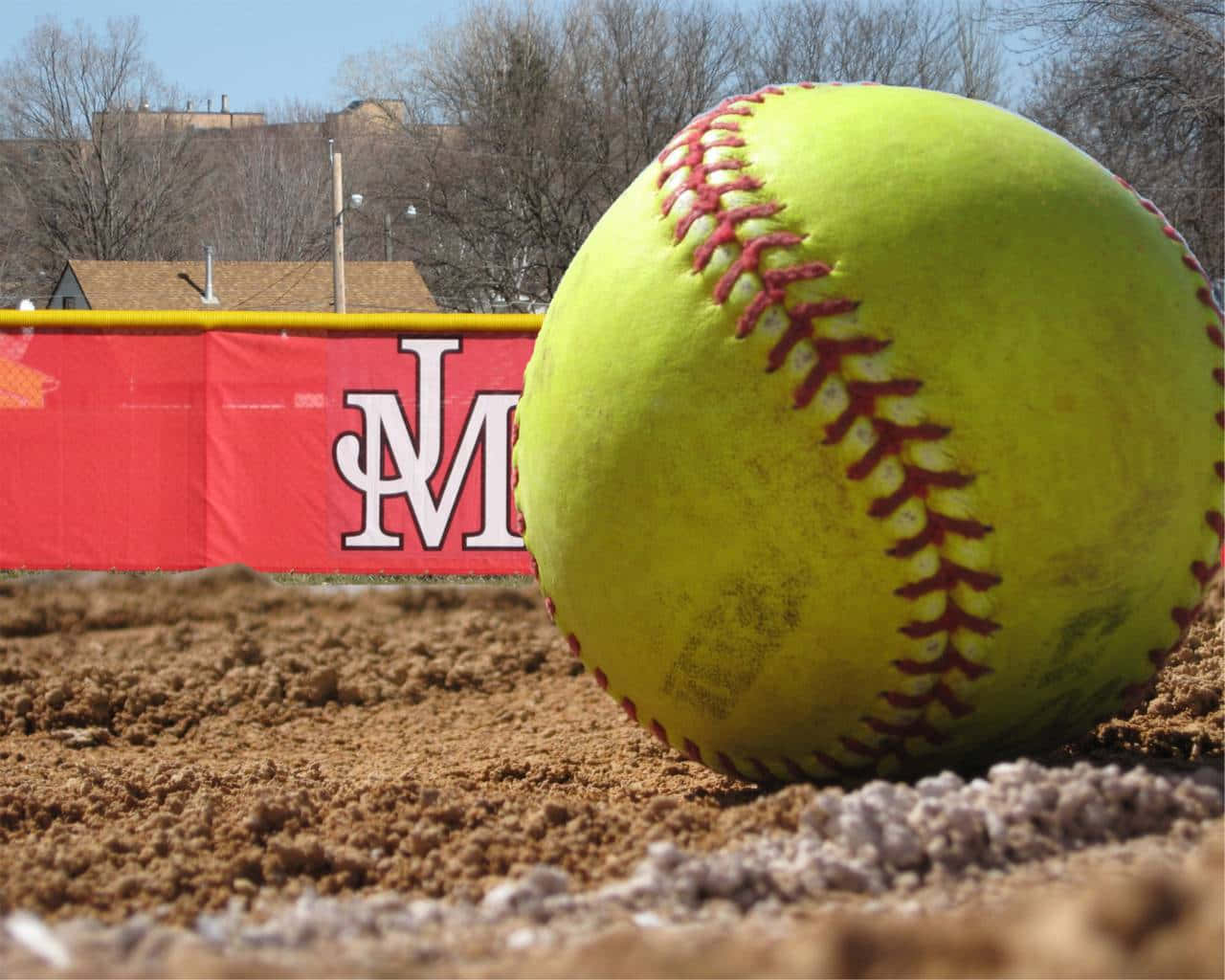 Softball 桌布 壁紙 — 一個鮮黃色帶紅色縫線的壘球放在棕色泥土上，背景有紅色橫幅和光禿禿的樹木。