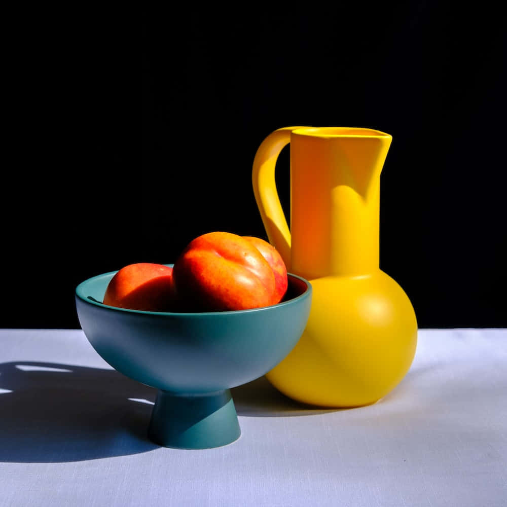 still life 桌布 壁紙 — 一個鮮黃色水壺和一個青綠色碗，碗裡放著桃子，在黑色背景下的白色布料上投下銳利的陰影。
