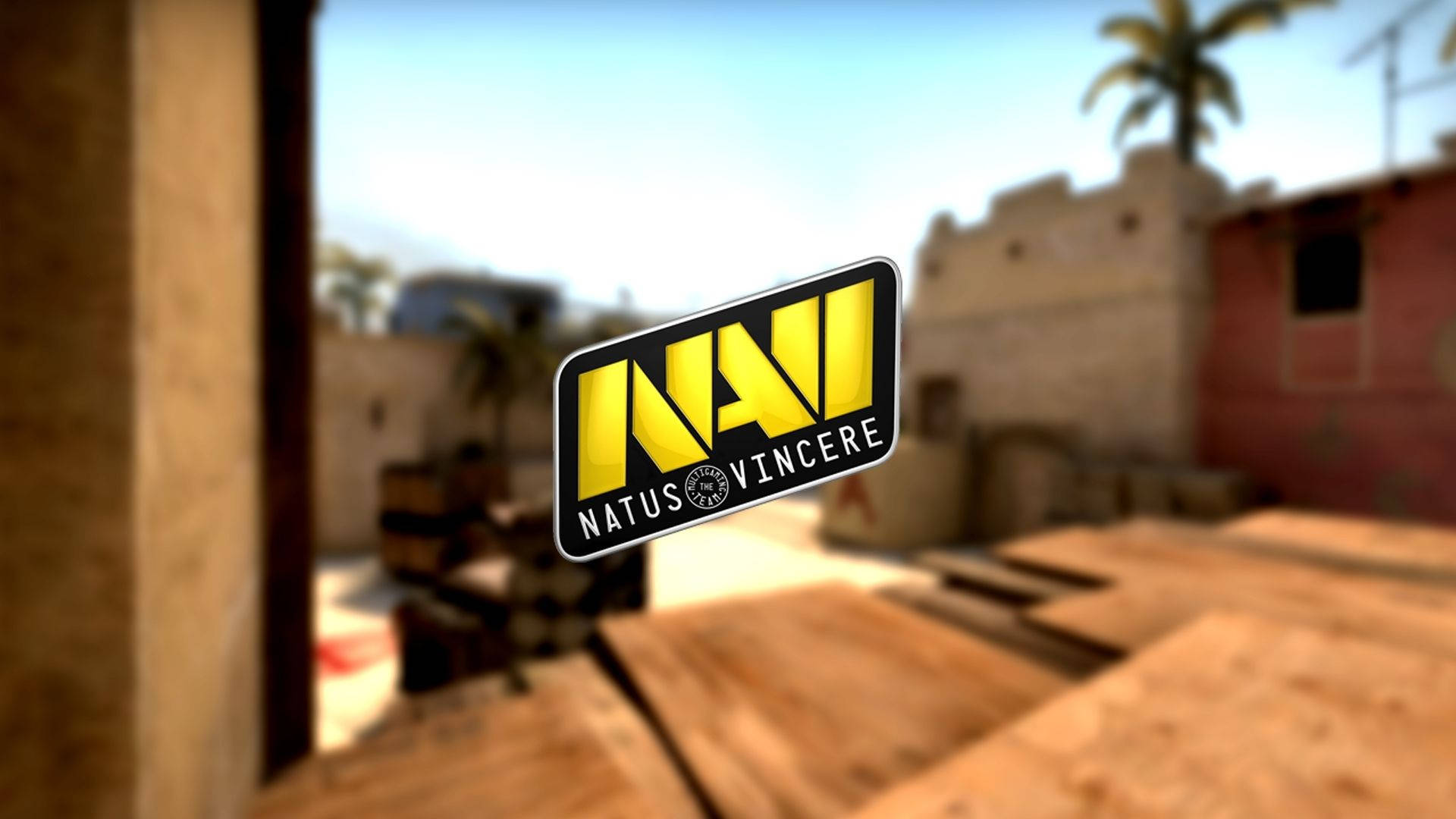 Natus Vincere 桌布 壁紙 — 一個鮮黃色的Natus Vincere標誌漂浮在模糊的沙漠主題遊戲地圖前，地圖上有木板和石頭建築。