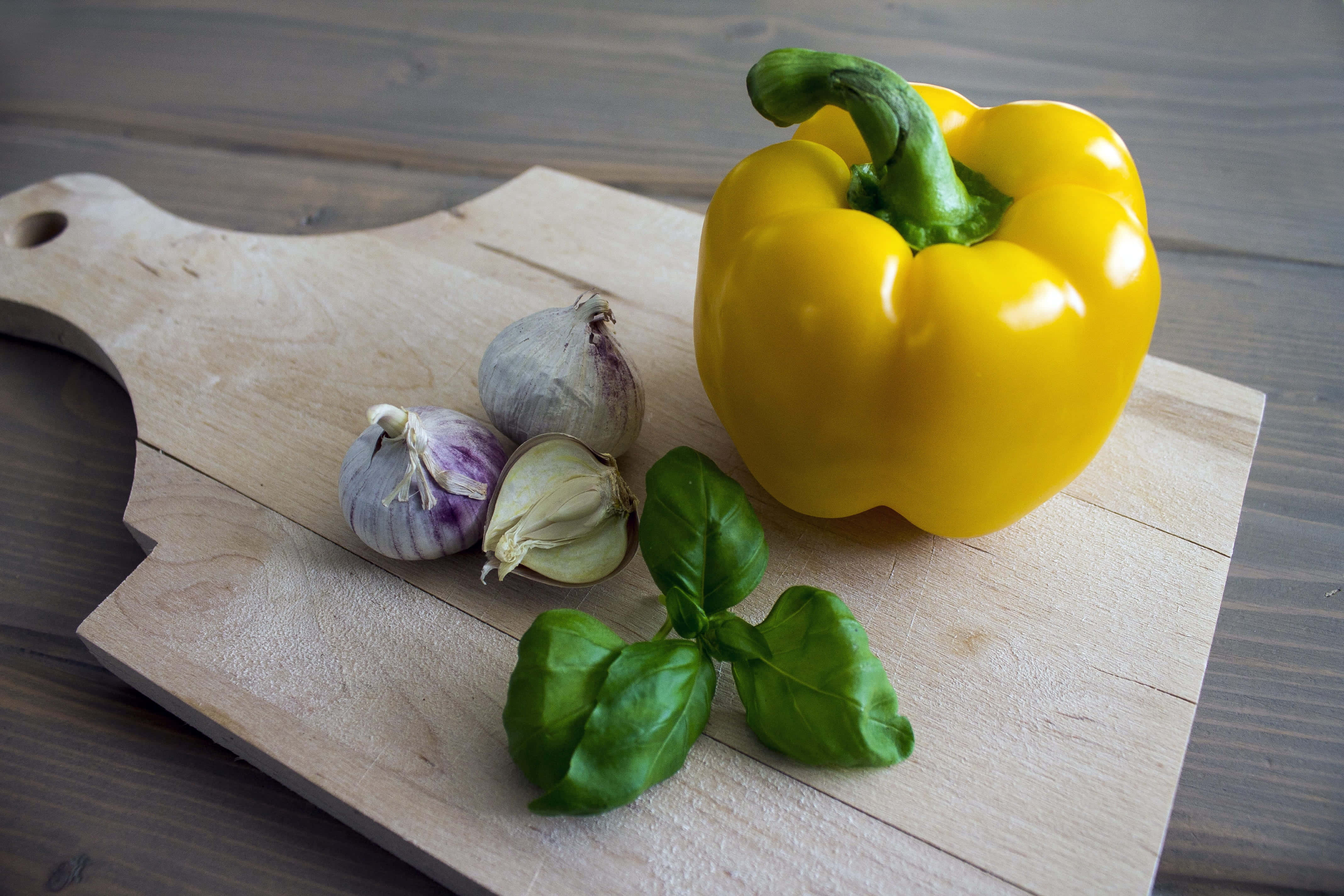 Yellow Bell Pepper 桌布 壁紙 — 一個鮮黃色的甜椒、蒜瓣和新鮮羅勒葉放在淺色木砧板上。