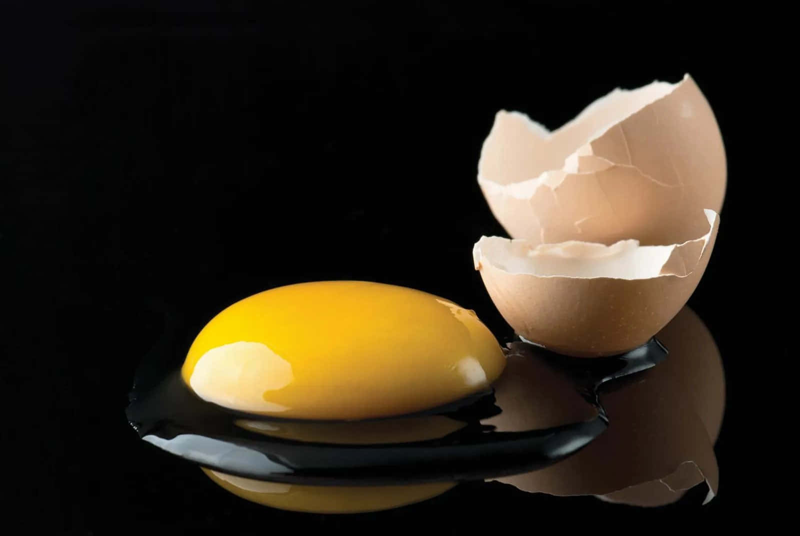 Egg 桌布 壁紙 — 一個鮮黃色的蛋黃放在黑色反光表面上，旁邊有一個破裂的棕色蛋殼，背景為深色。