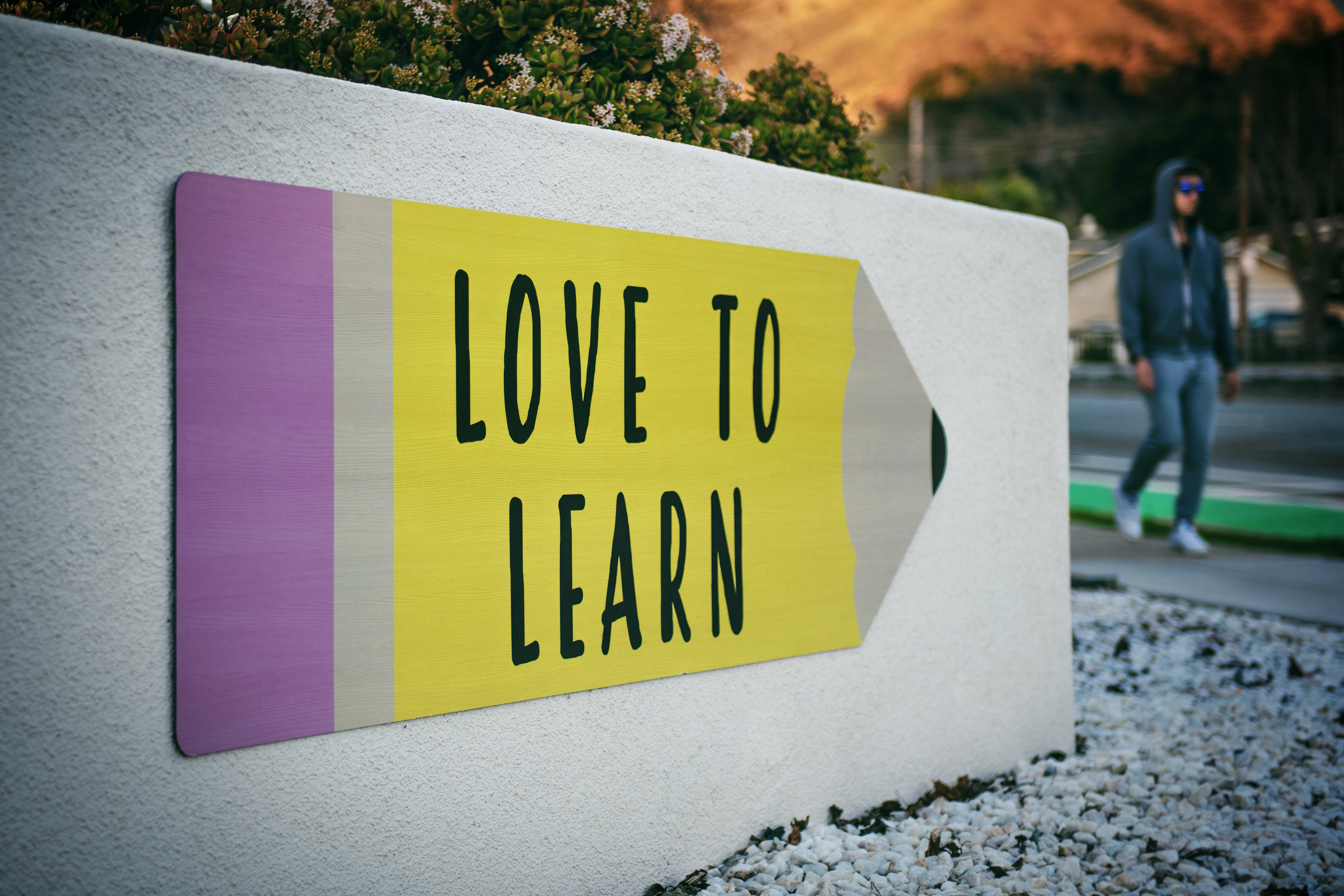 the more you know 桌布 壁紙 — 一個鮮黃色鉛筆形狀的標牌，上面寫著「LOVE TO LEARN」，安裝在白色牆壁上，背景中有一個人走過。