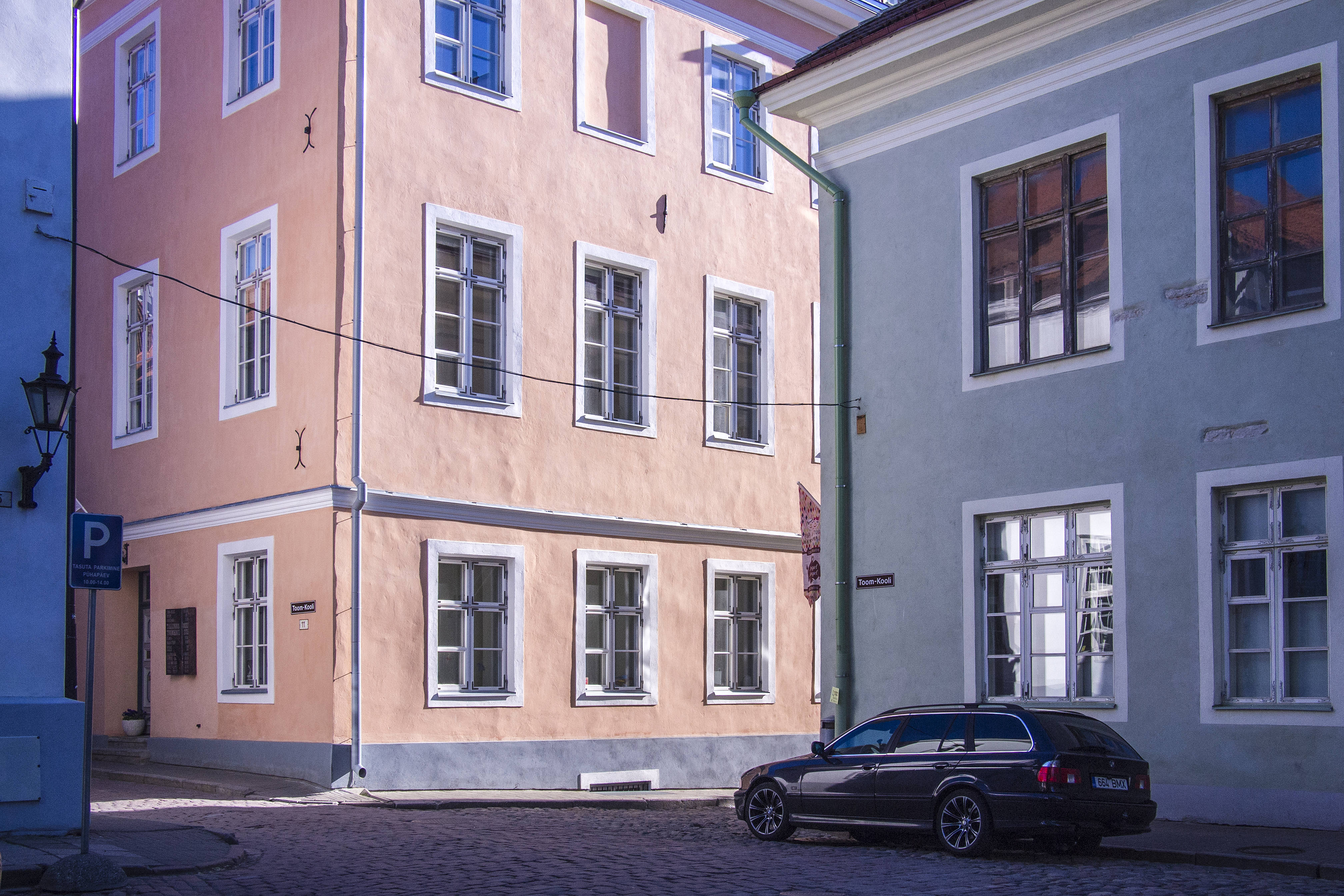 Estonia 桌布 壁紙 — 一個鵝卵石街道的角落，有粉彩色的建築物和一輛深色汽車在明亮的日光下停泊。