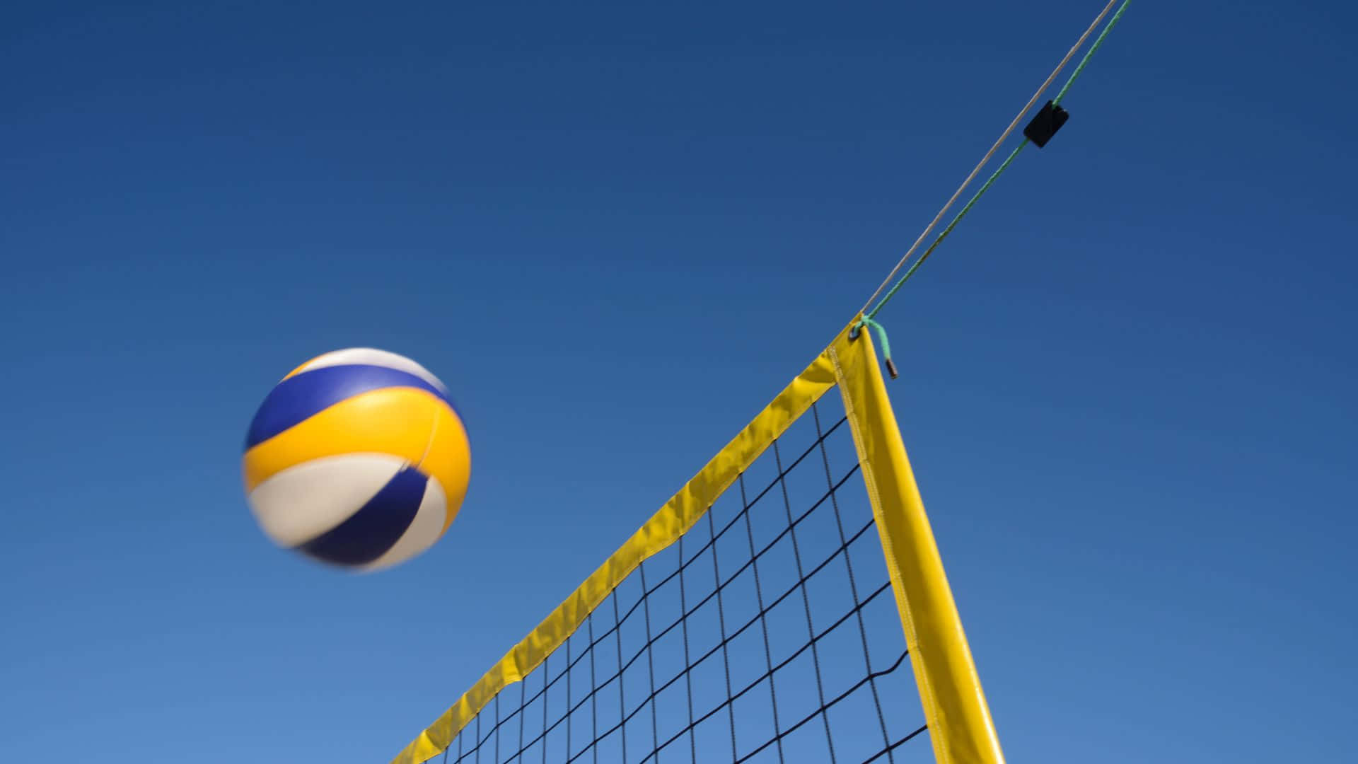 Best Sports 桌布 壁紙 — 一個黃、藍、白色的排球在空中模糊，正接近一個明亮的黃色球網，背景是清澈的藍天。