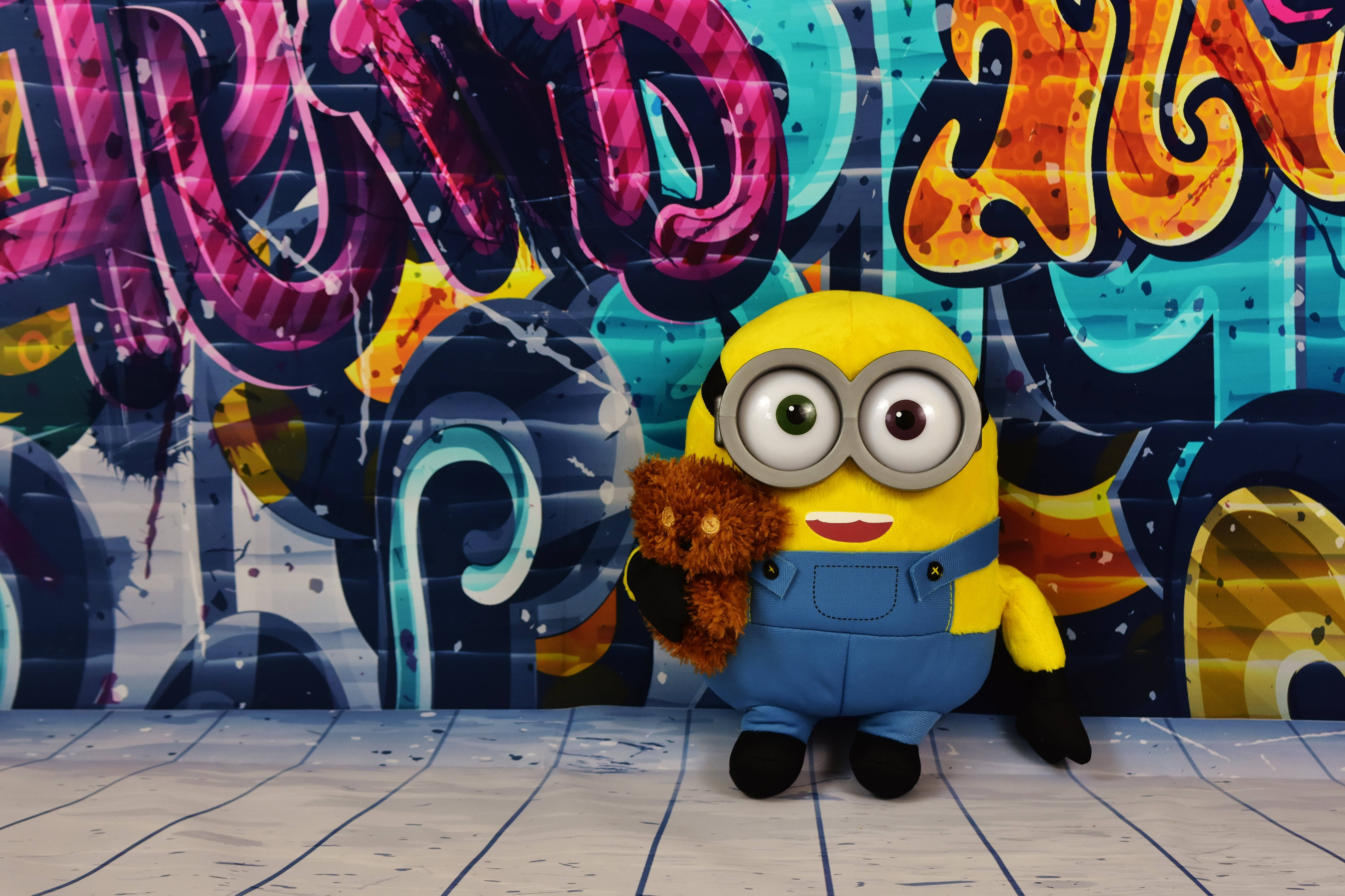 小黃人 桌布 壁紙 — 一個黃色Minion毛絨玩具抱著一個棕色泰迪熊，站在充滿活力的塗鴉牆前。
