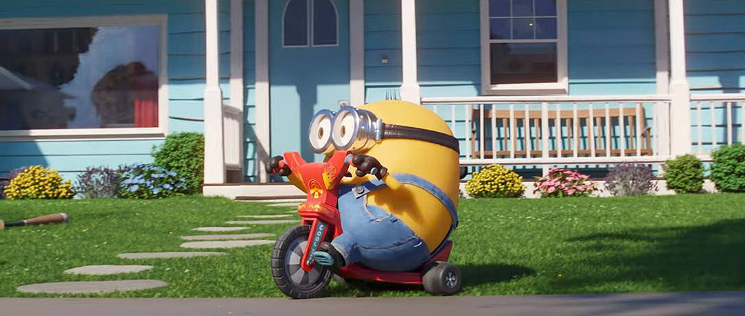 gru 桌布 壁紙 — 一個黃色Minion穿著牛仔短褲，在陽光明媚的草坪上騎著紅色三輪車，前面是一棟藍色白邊的房子。