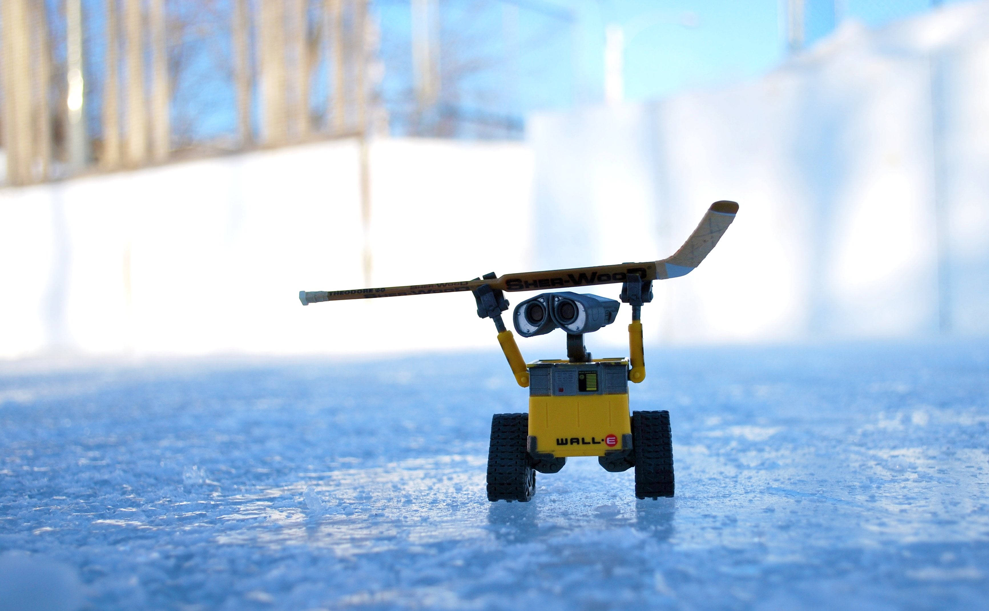 hockey 桌布 壁紙 — 一個黃色WALL-E玩具機器人站在冰藍色的地面上，在明亮的冬日天空下拿著一支曲棍球棍。