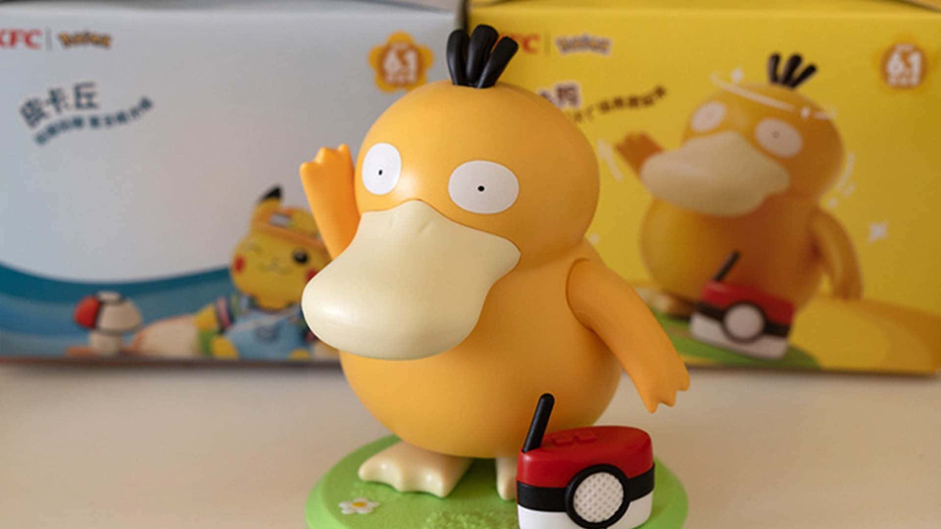 Pokemon Figures 桌布 壁紙 — 一個黃色卡通鴨子寶可夢公仔在綠色底座上，在精靈球旁邊開心地揮手，後面有模糊的產品盒。