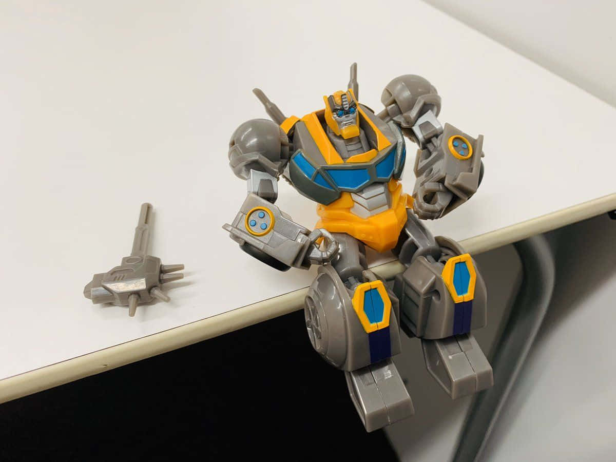 Animated 桌布 壁紙 — 一個黃色和灰色的Bumblebee變形金剛玩具機器人坐在白色書桌邊緣，其分離的武器在附近。