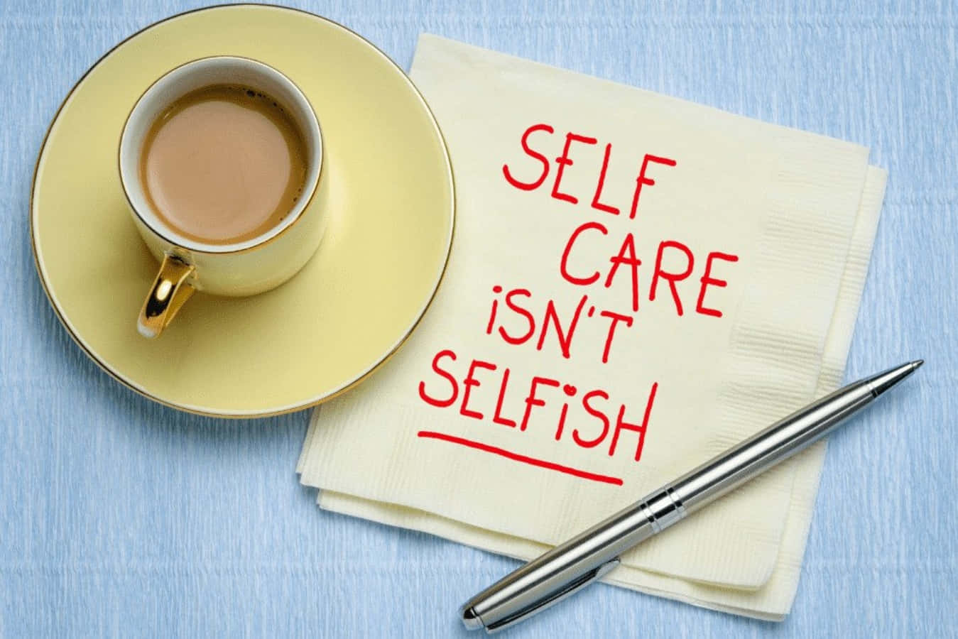 Self Care 桌布 壁紙 — 一個黃色咖啡杯放在藍色紋理表面上，旁邊有一張紅色手寫文字寫著「SELF CARE ISN'T SELFISH」的餐巾和一支銀色筆。