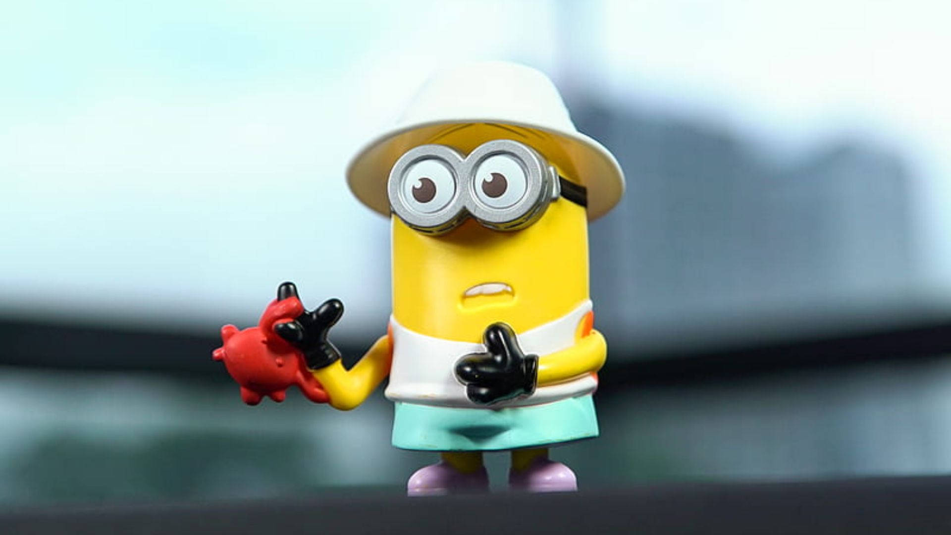 minions the rise of gru 桌布 壁紙 — 一個黃色小小兵玩具，戴著白色帽子和護目鏡，站在深色表面上，手持一隻紅色螃蟹。