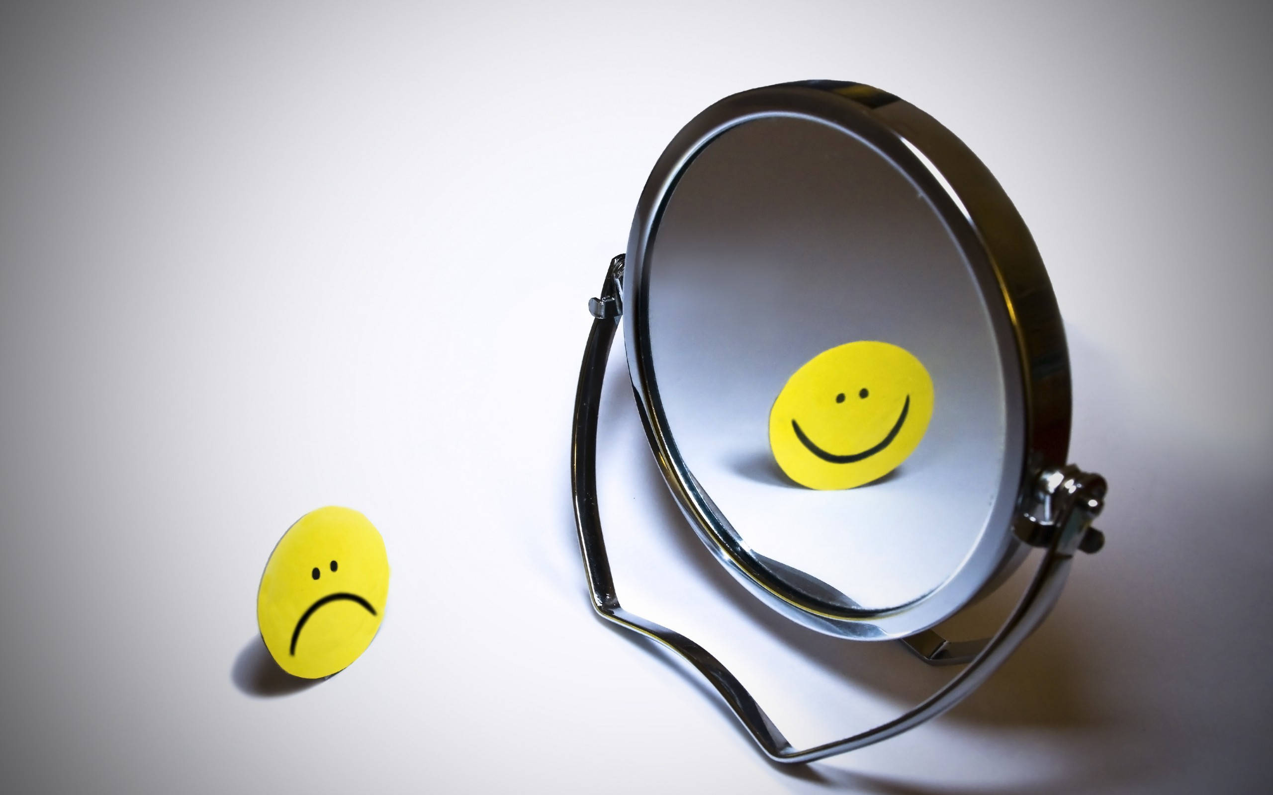 Smile 桌布 壁紙 — 一個黃色悲傷表情貼紙與其在白色平面上的小圓鏡中的快樂倒影形成對比。