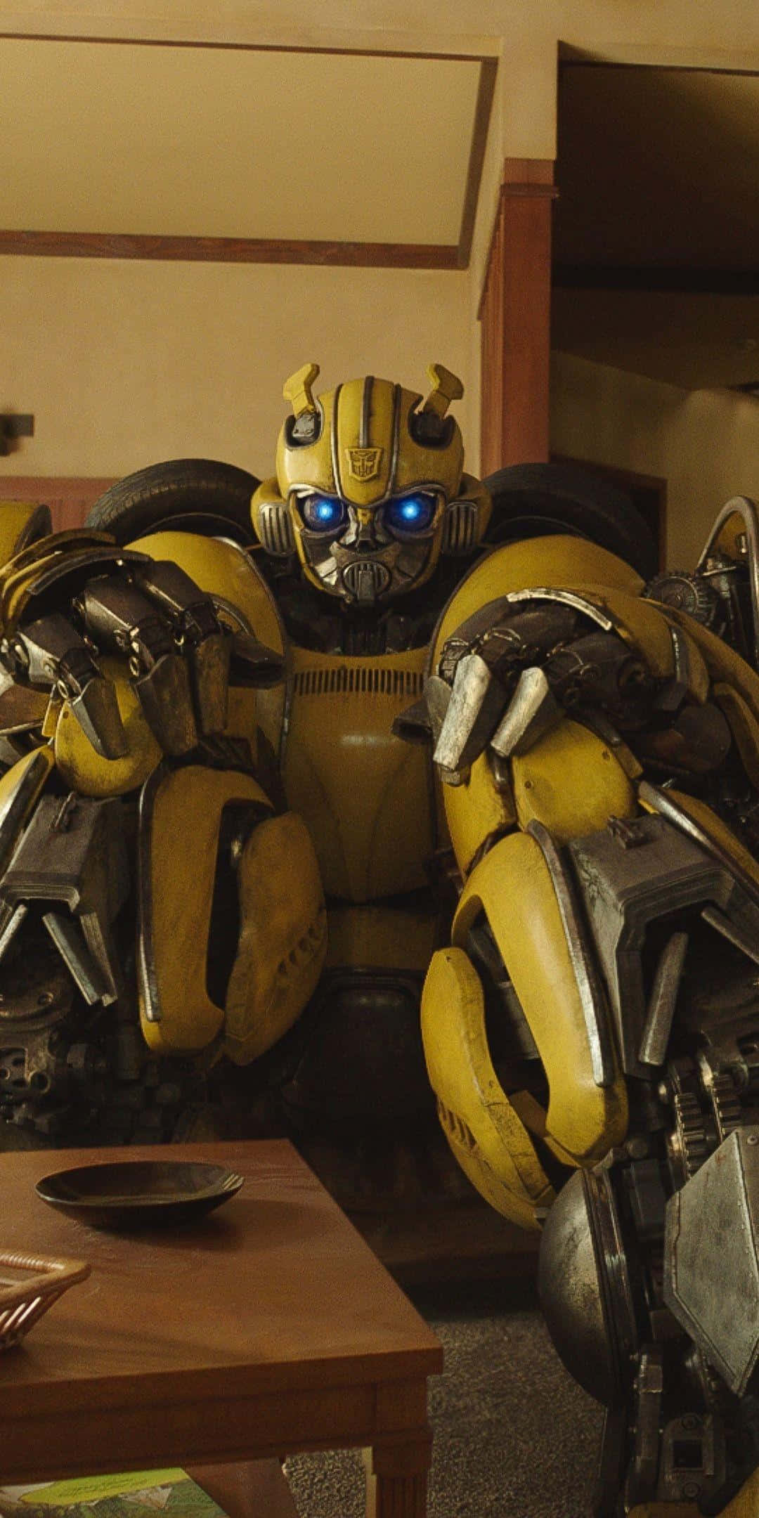 Bumblebee Transformer 桌布 壁紙 — 一個黃色機器人，有著發光的藍色眼睛，坐在一個有桌子和碗的木製房間裡。