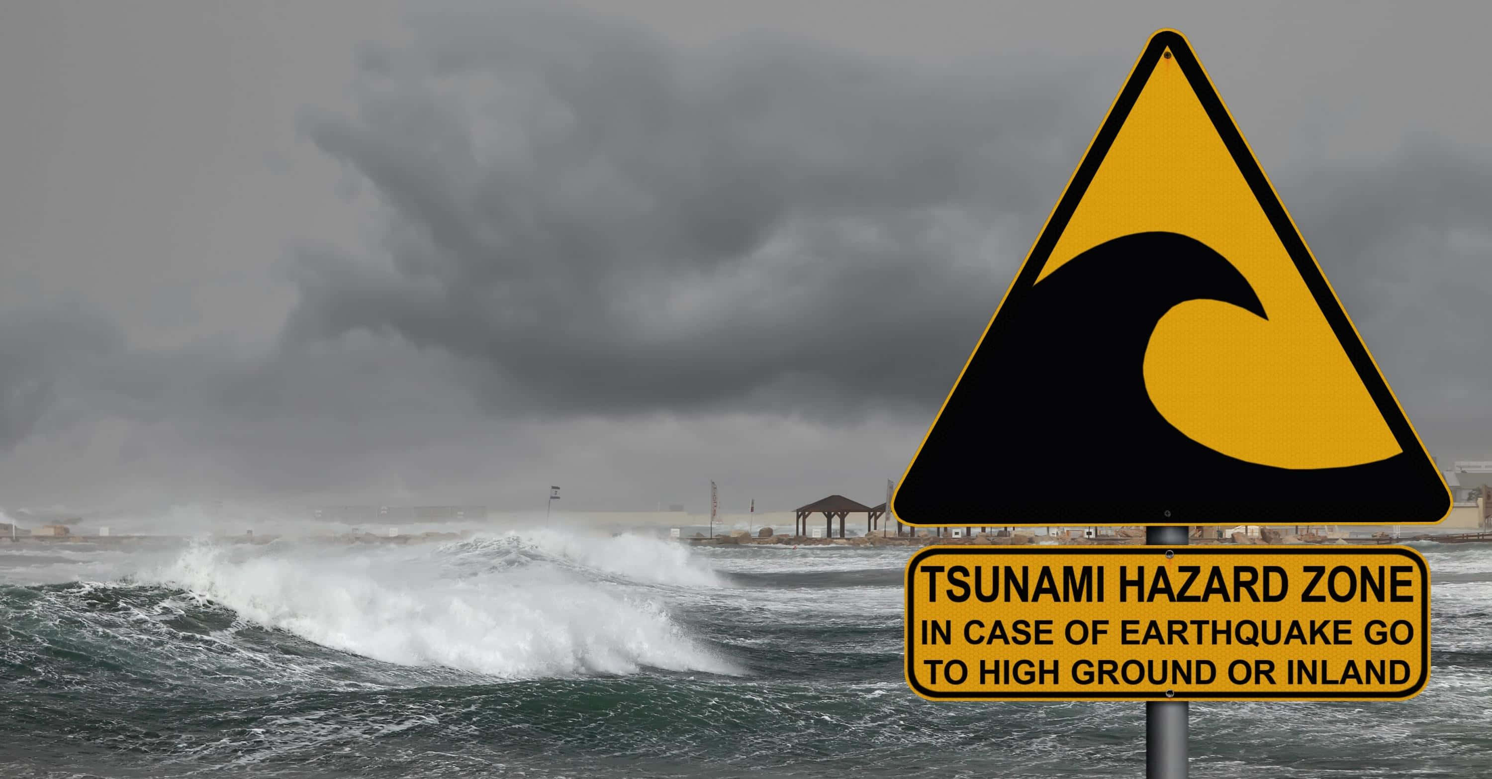 Tsunami 桌布 壁紙 — 一個黃色海嘯警告標誌聳立在沙灘附近，背景是翻騰的黑暗海浪和暴風雨的天空。