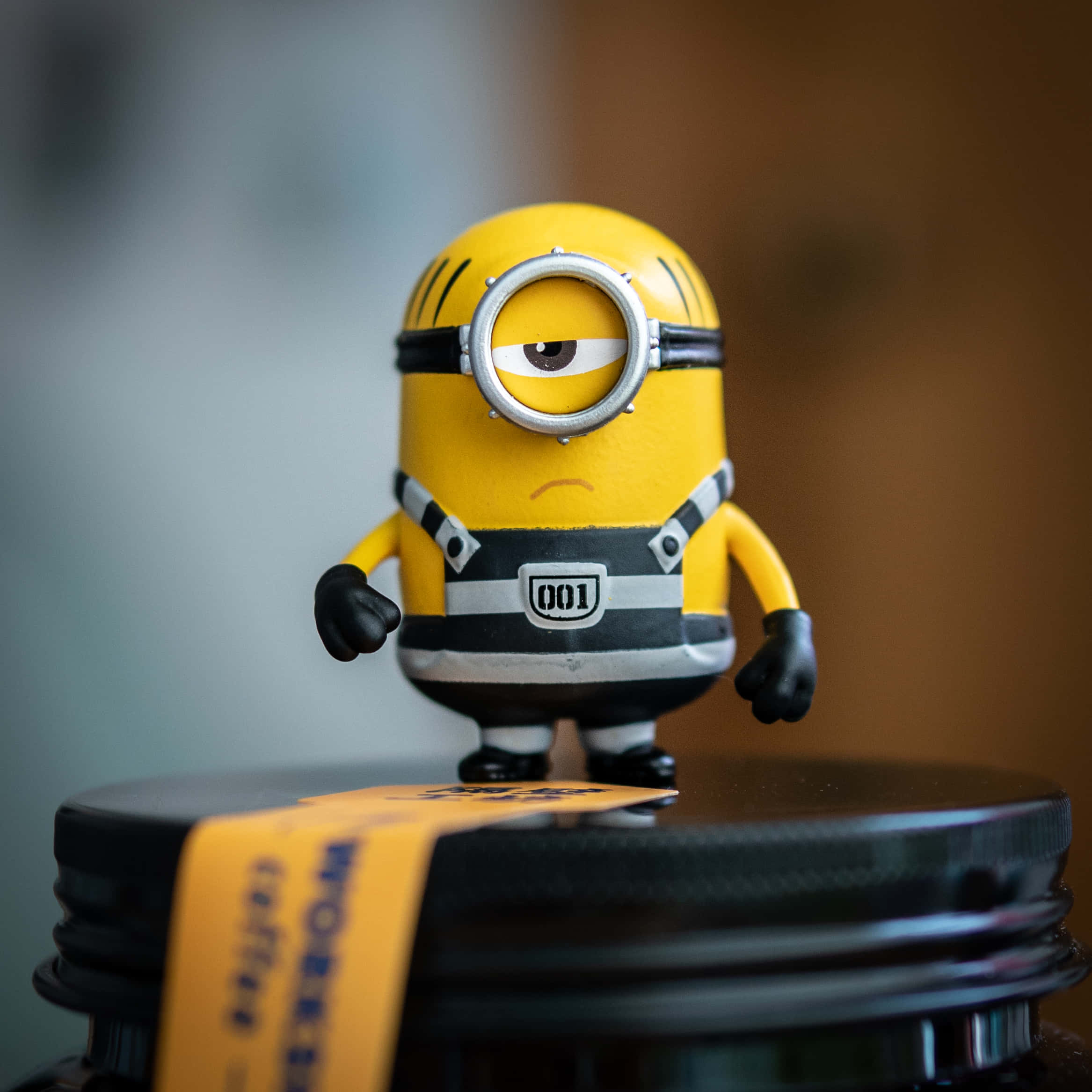 小黃人 桌布 壁紙 — 一個黃色的Minion公仔，有一隻眼睛，戴著'001'徽章，站在一個黑色的罐子上，罐子有黃色標籤。