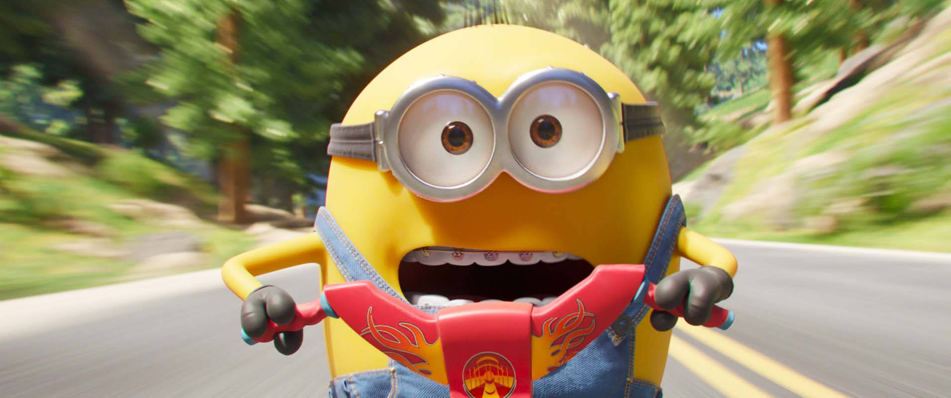 gru 桌布 壁紙 — 一個黃色的Minion戴著護目鏡和牛仔吊帶褲，開心地騎著紅色滑板車在森林道路上行駛。