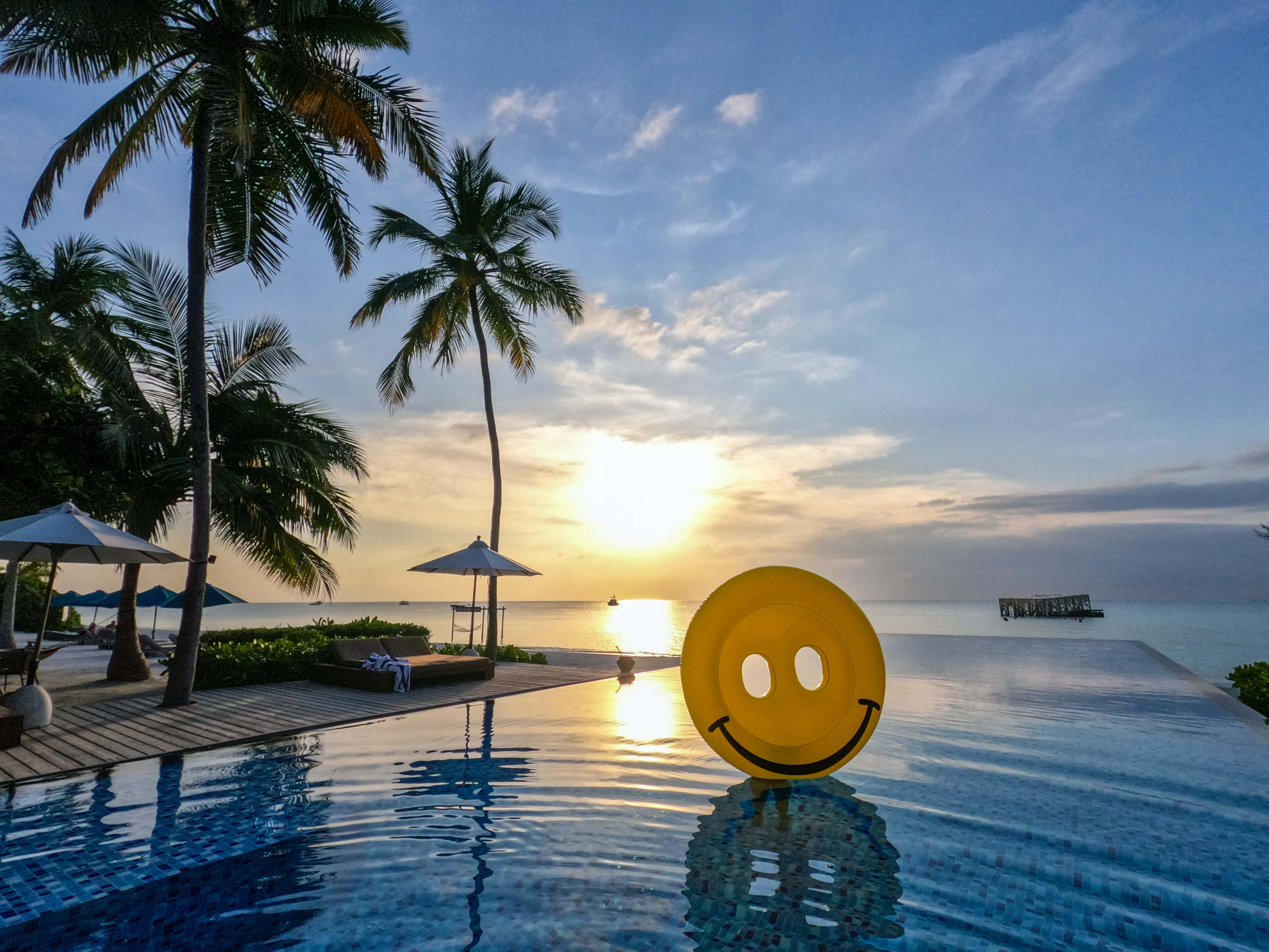 Summer Vacation 桌布 壁紙 — 一個黃色笑臉浮床在無邊際泳池中漂浮，夕陽在棕櫚樹和海洋後方落下。