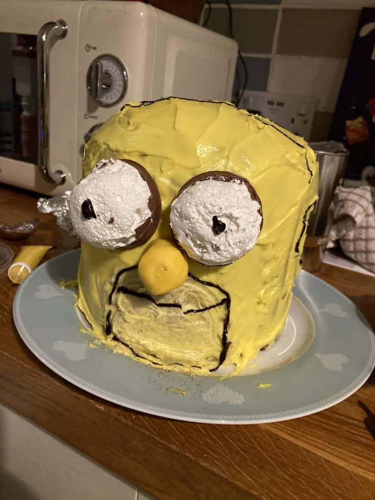 Birthday Cake 桌布 壁紙 — 一個黃色糖霜蛋糕，造型像小小兵，有巧克力眼睛和檸檬鼻子，放在廚房的盤子上。