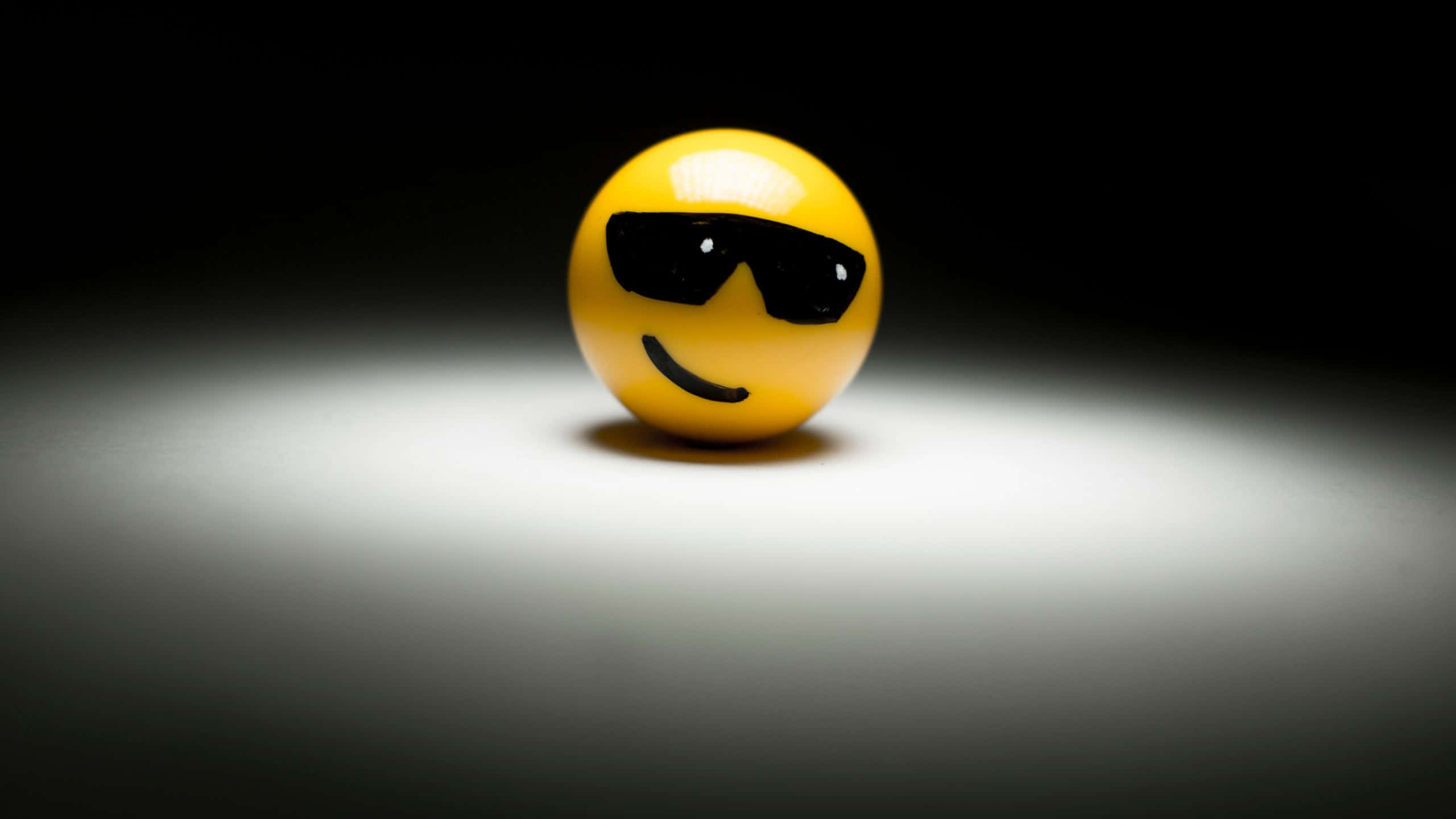 Smile Emoji 桌布 壁紙 — 一個黃色表情符號球戴著黑色太陽眼鏡，坐在黑暗漸層地板上的聚光燈下中央位置。