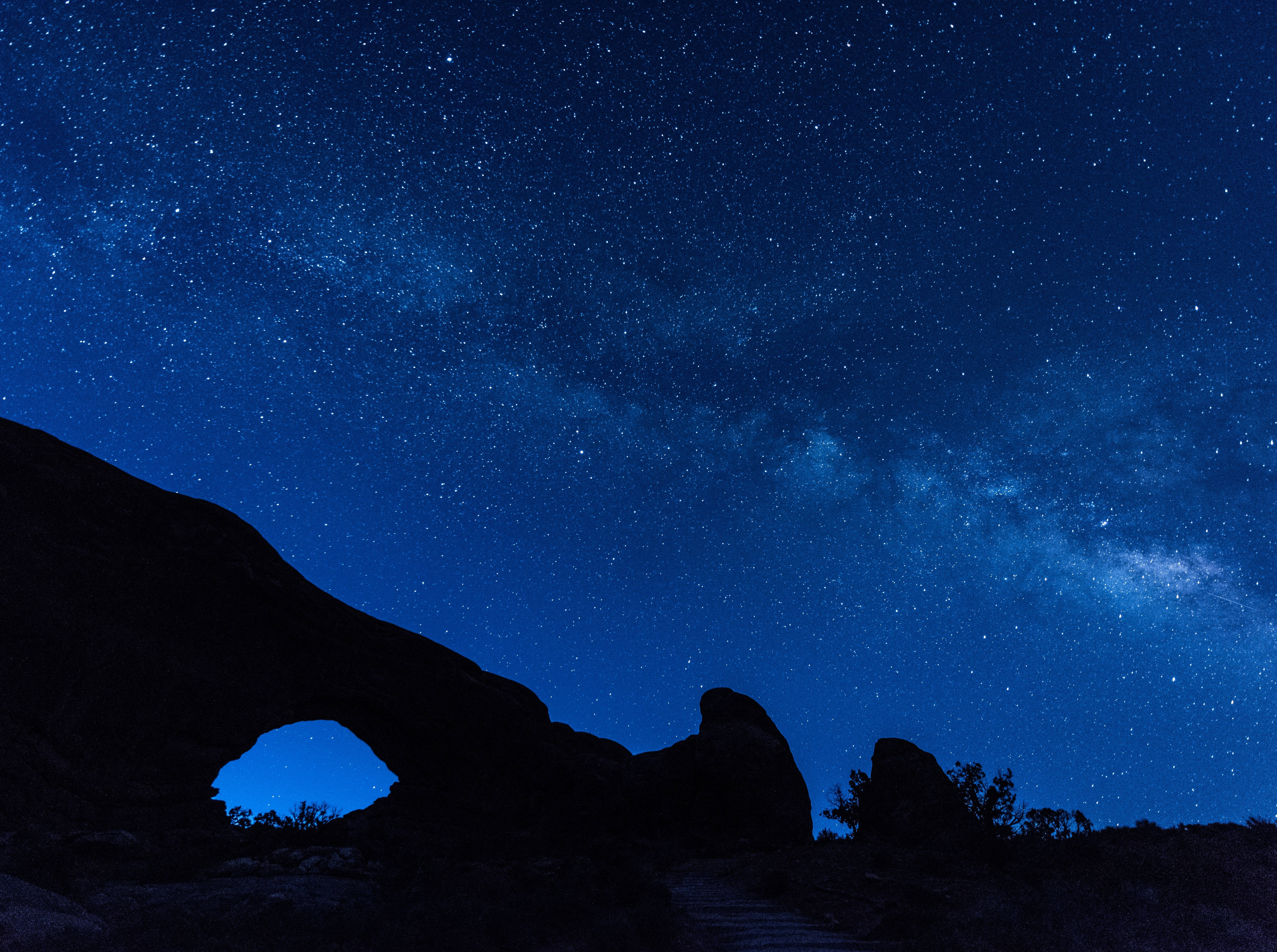夜空 桌布 壁紙 — 一個黑暗的岩石拱門框住了一個充滿無數星星和銀河系發光帶的發光藍色天空。