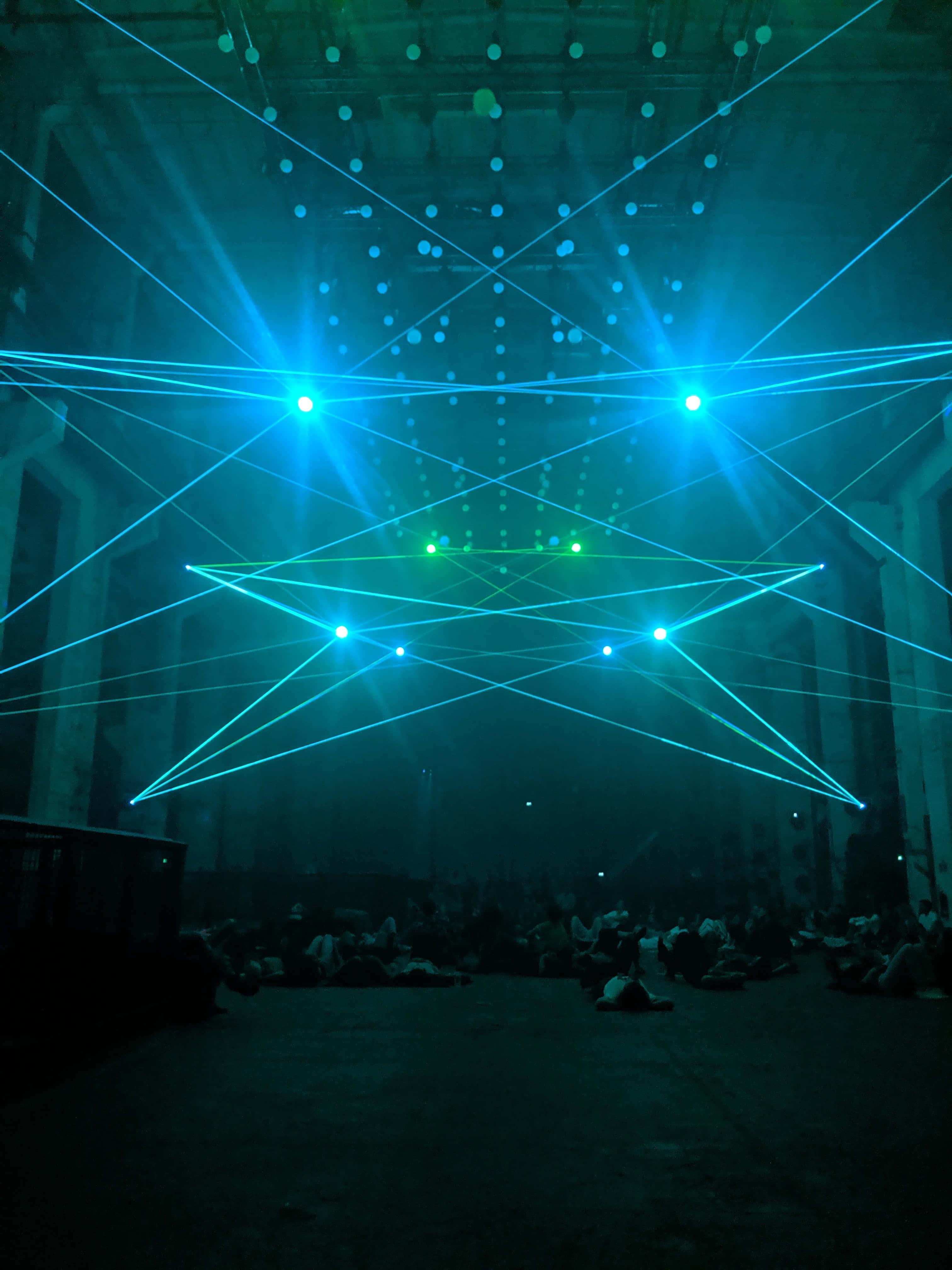 Laser 桌布 壁紙 — 一個黑暗的工業大廳，充滿了交叉的藍色和綠色雷射光束，在坐著的觀眾上方形成幾何圖案。