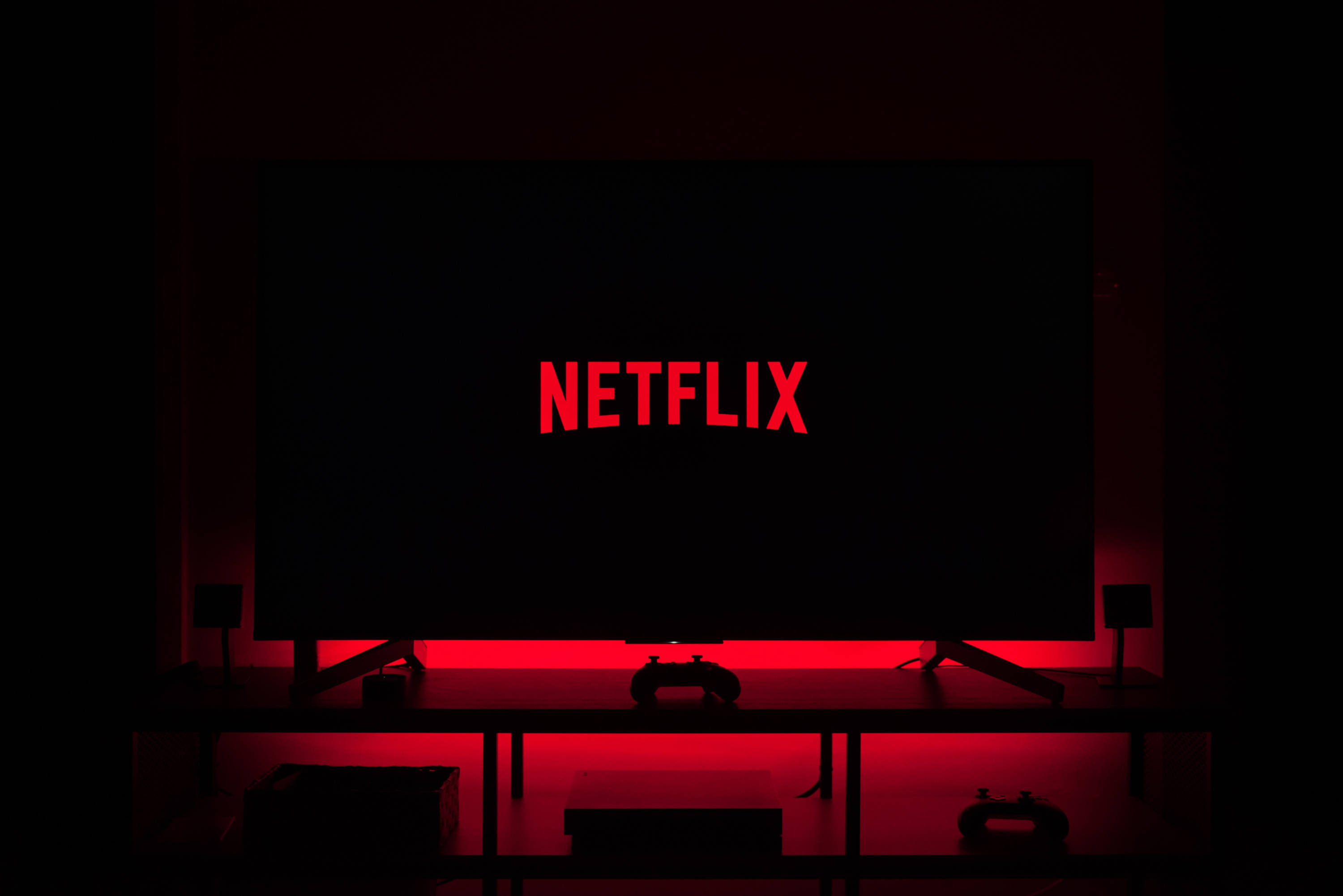 Netflix 桌布 壁紙 — 一個黑暗的房間，被顯示Netflix標誌的電視螢幕發出的紅光照亮，下方支架上有遊戲控制器。
