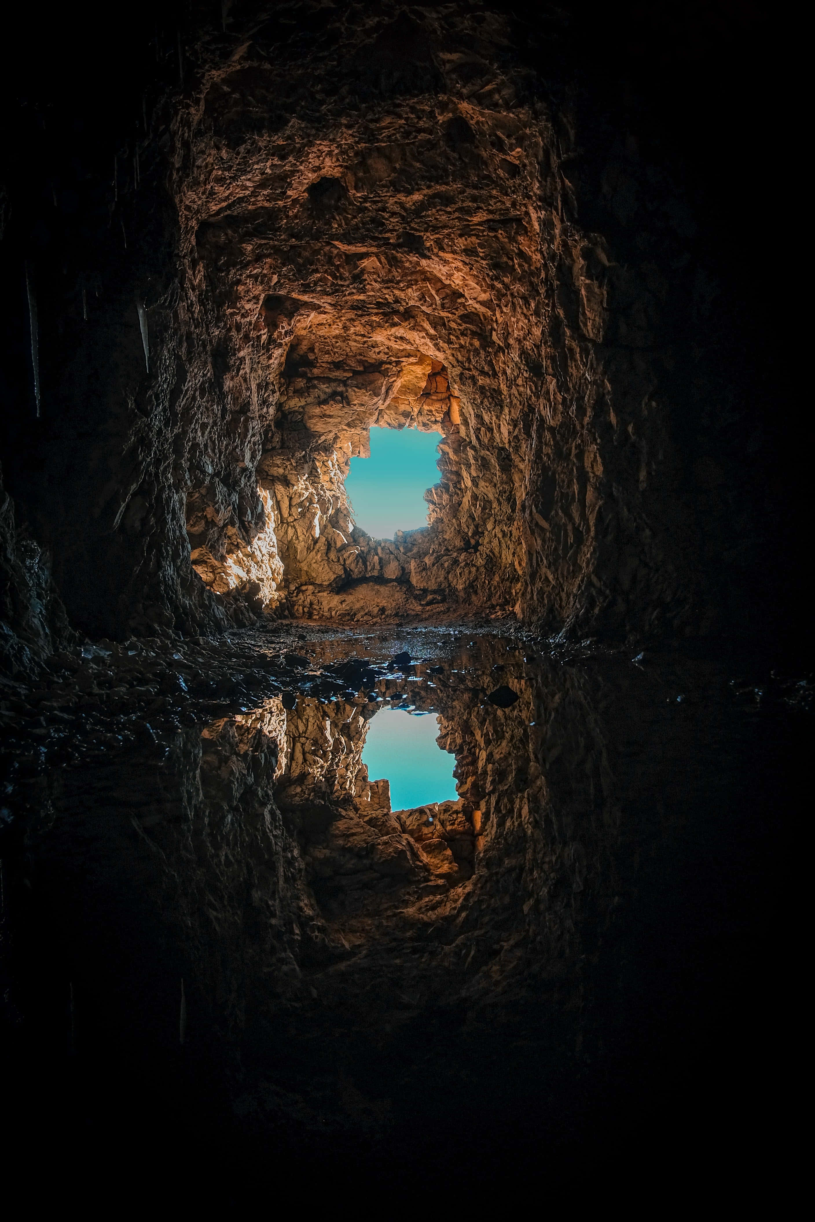 Cave 桌布 壁紙 — 一個黑暗的洞穴，粗糙的石牆框住了一片明亮的藍天，完美地倒映在下方平靜的水池中。