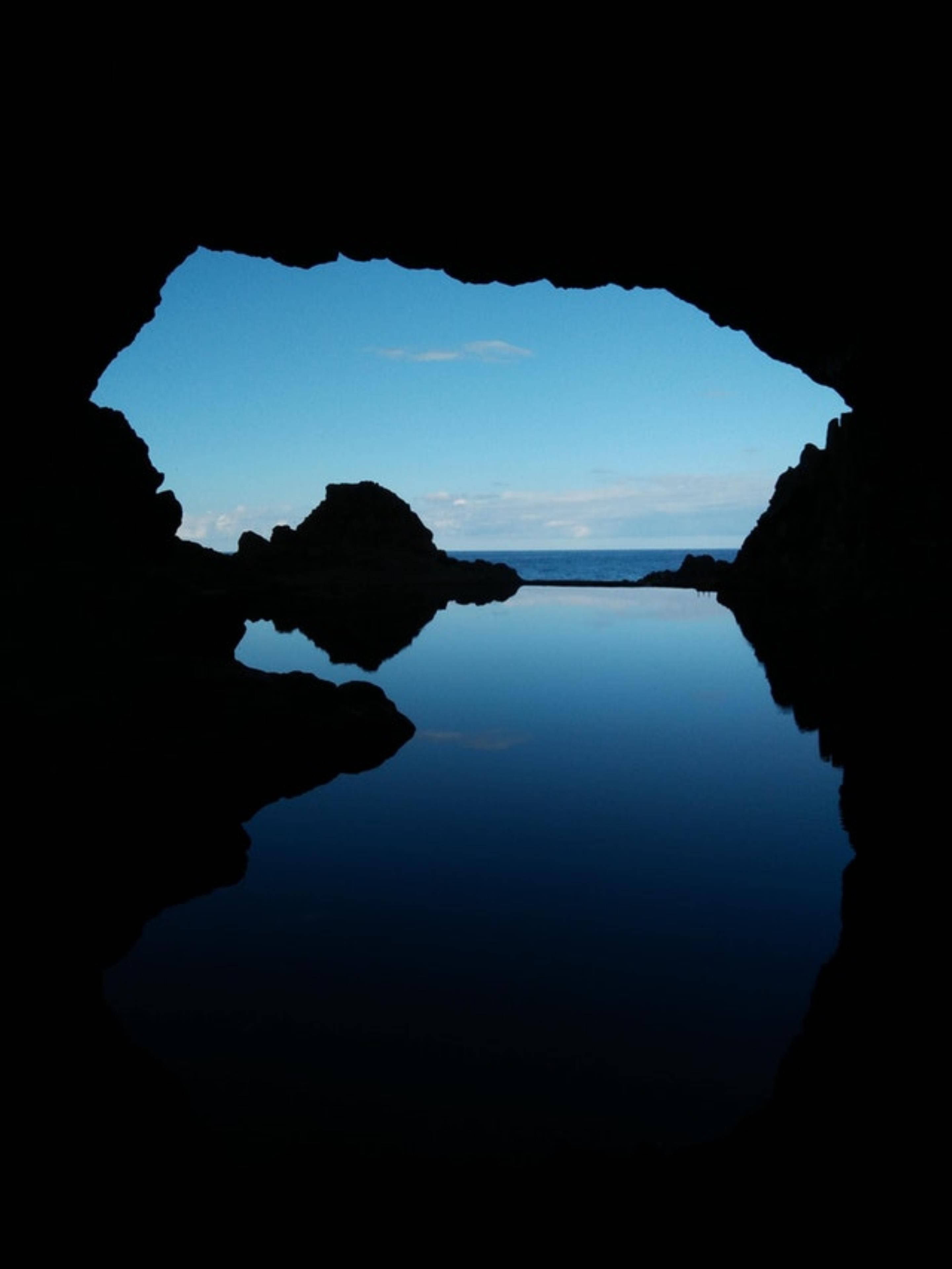 AMOLED 桌布 壁紙 — 一個黑暗的洞穴框住了一片寧靜的藍天和寧靜的海洋，完美地倒映在下面的靜水中。