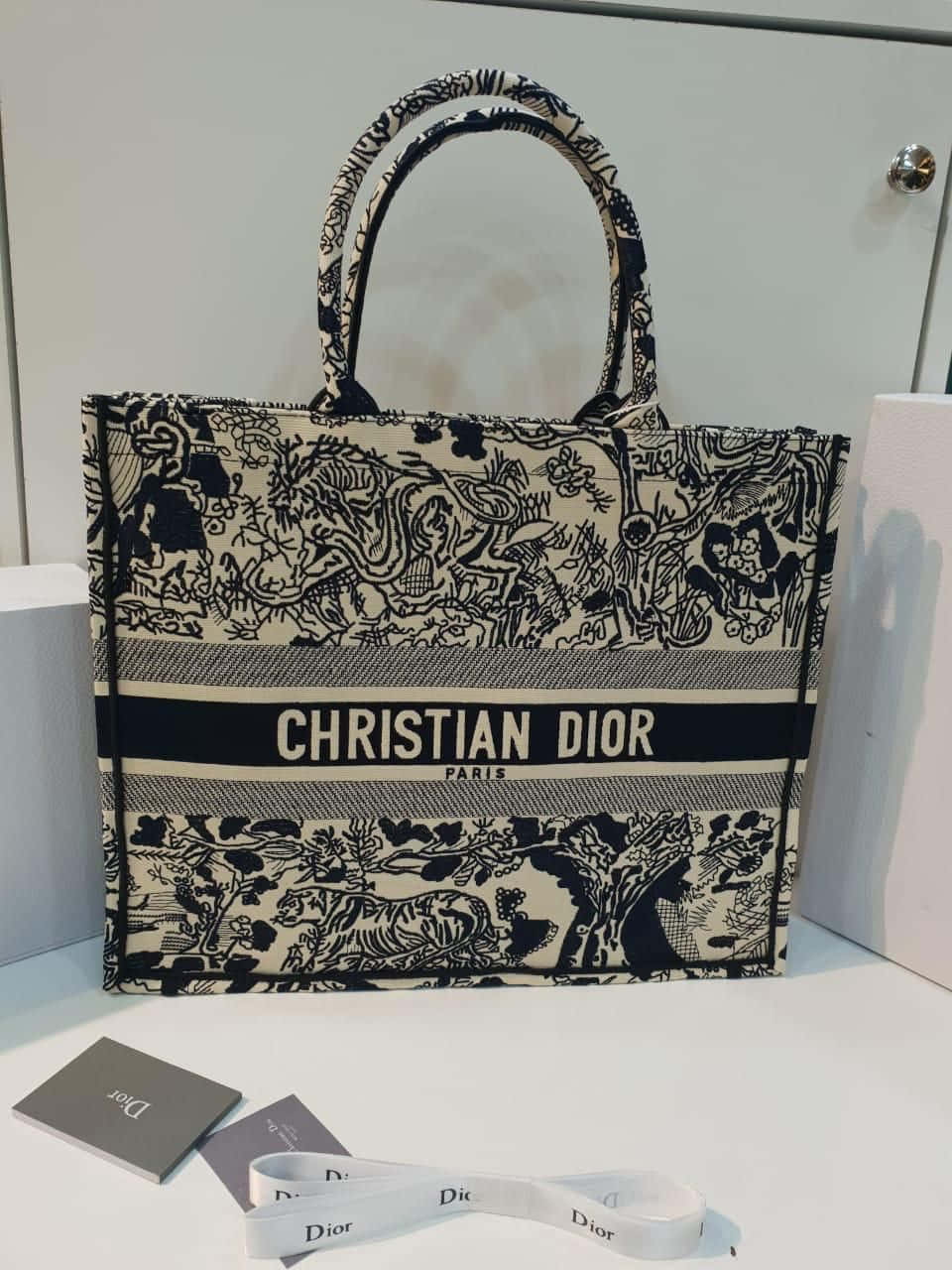 Dior 動態 桌布 壁紙 — 一個黑白Christian Dior托特包，帶有複雜的叢林印花，放在白色表面上，旁邊是品牌配件。