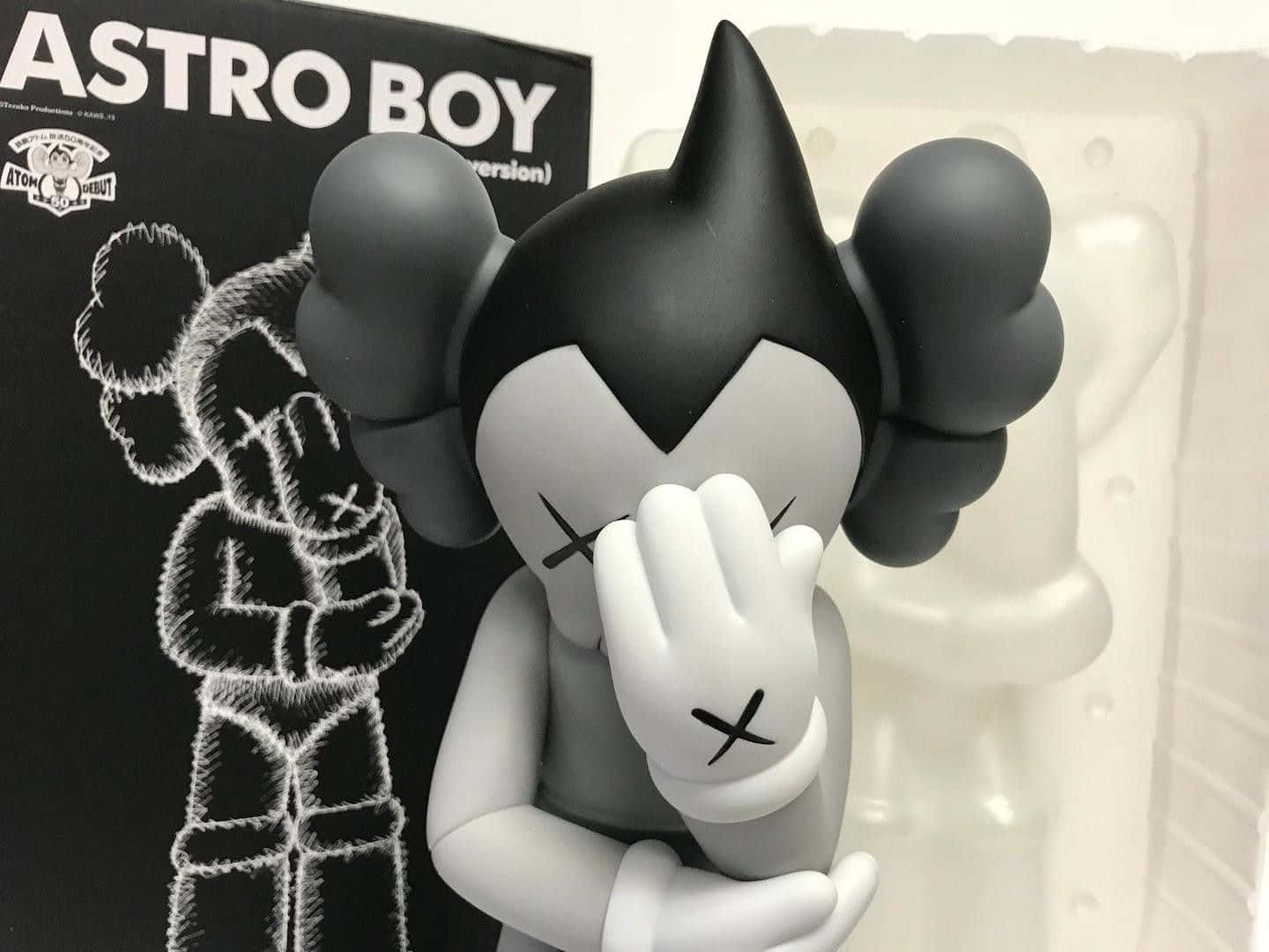 Kaws Astro Boy 桌布 壁紙 — 一個黑白KAWS Astro Boy搪膠公仔用一隻帶有X標記的手遮住眼睛，站在它的盒子旁邊。