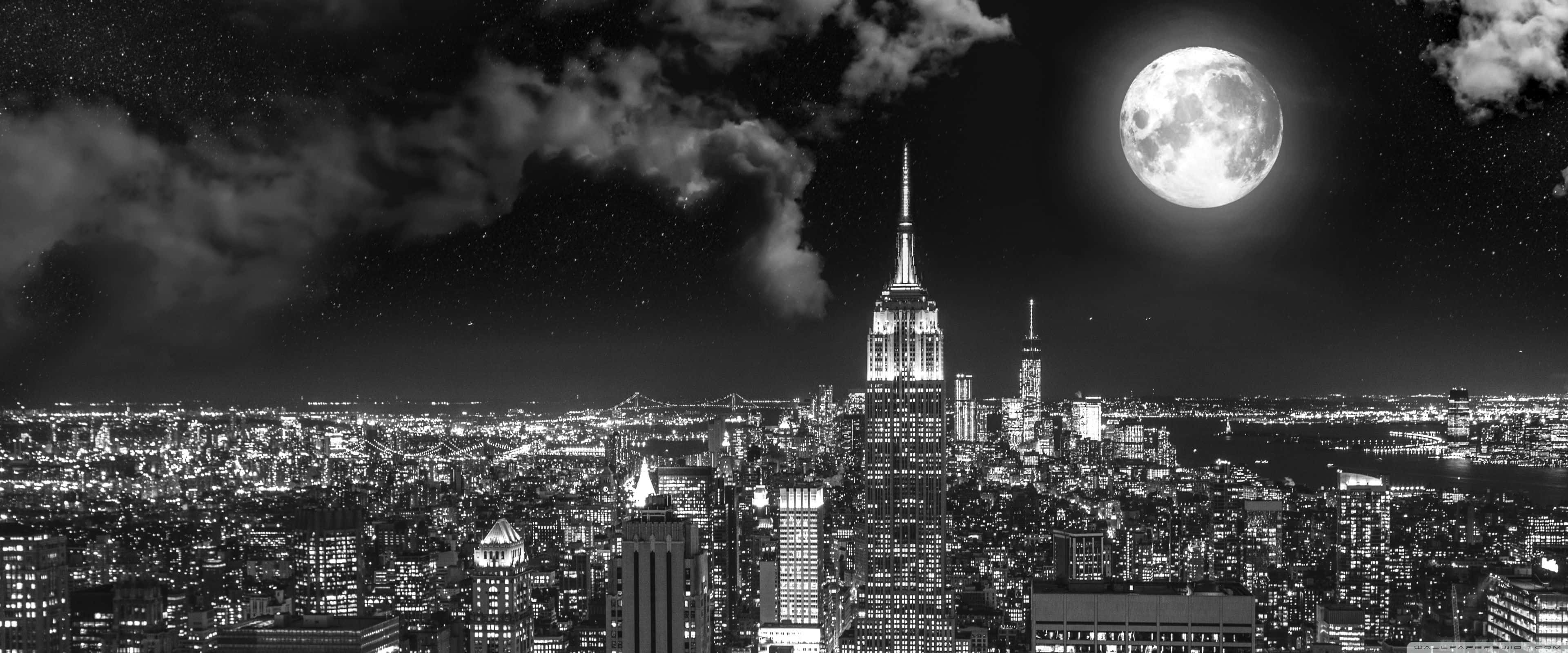 Apple 3840x1600 桌布 壁紙 — 一個黑白全景視圖，顯示夜晚城市天際線，下方是巨大的滿月，伴有零星雲朵。