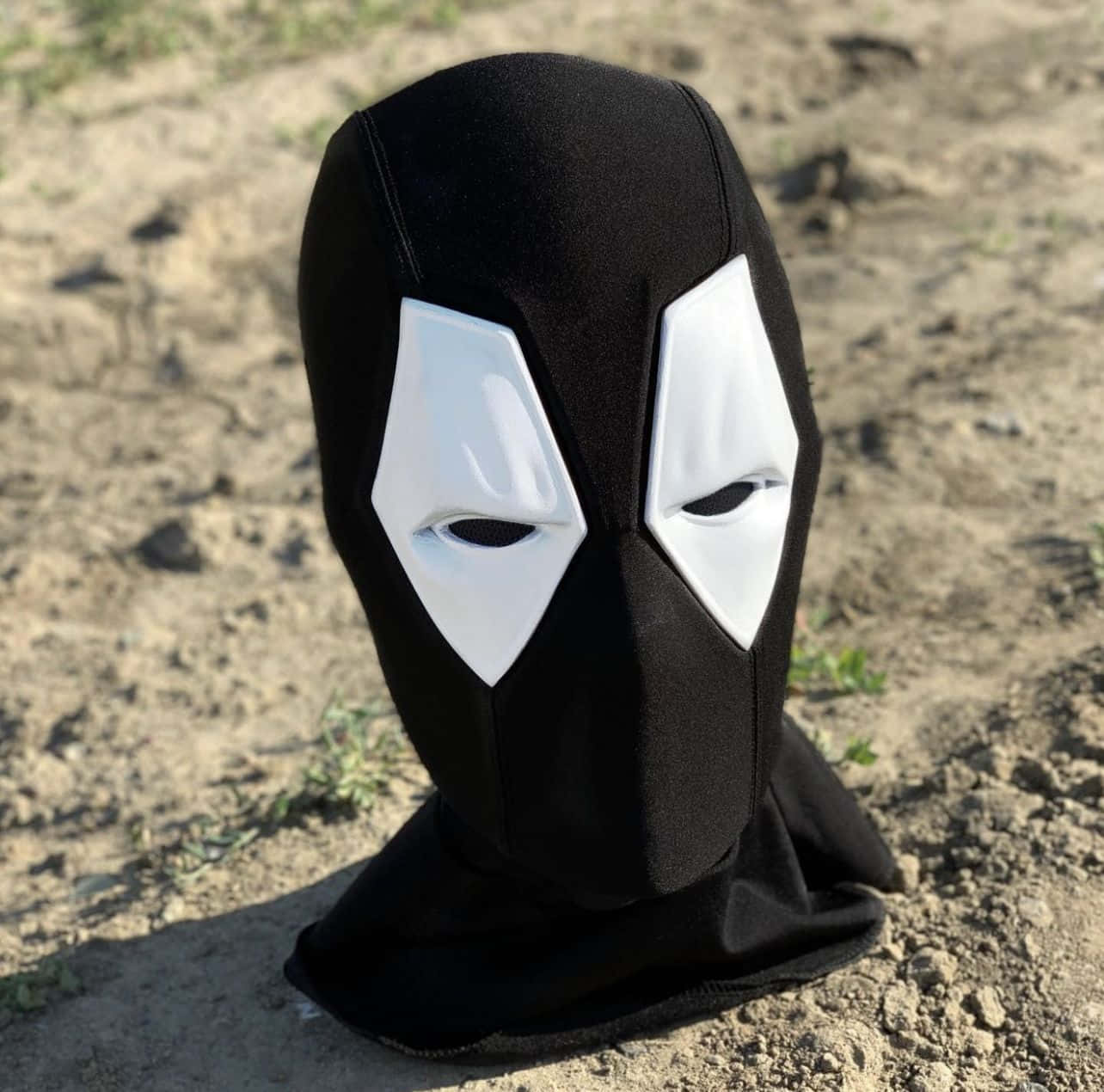 Deadpool Mask 桌布 壁紙 — 一個黑白死侍巴拉克拉瓦面具在明亮日光下放在乾燥泥土上。