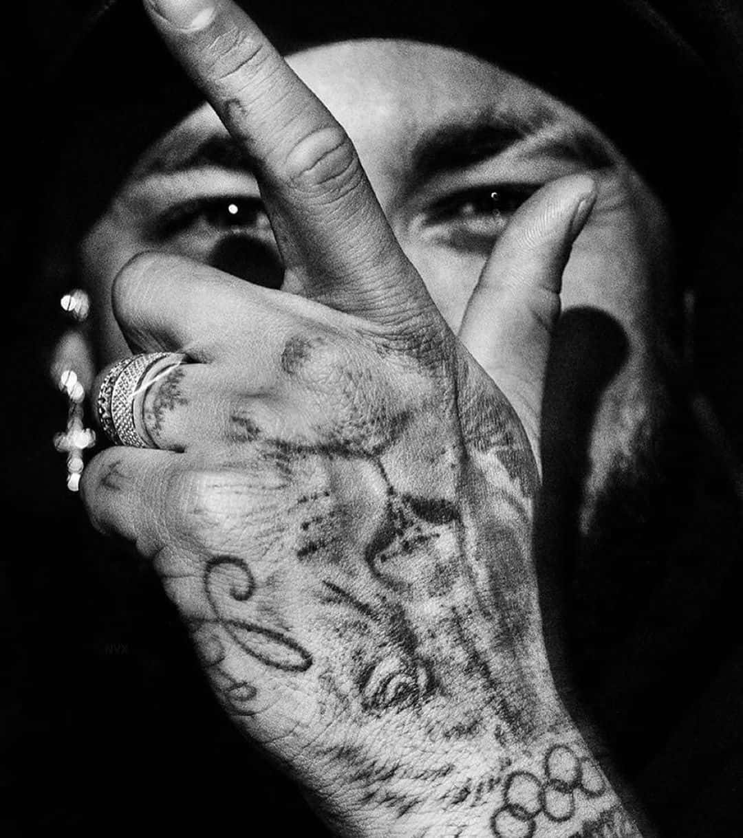 Tattoo 桌布 壁紙 — 一個黑白特寫，一隻有刺青的手在一個眼神強烈的男人臉上做出和平手勢。