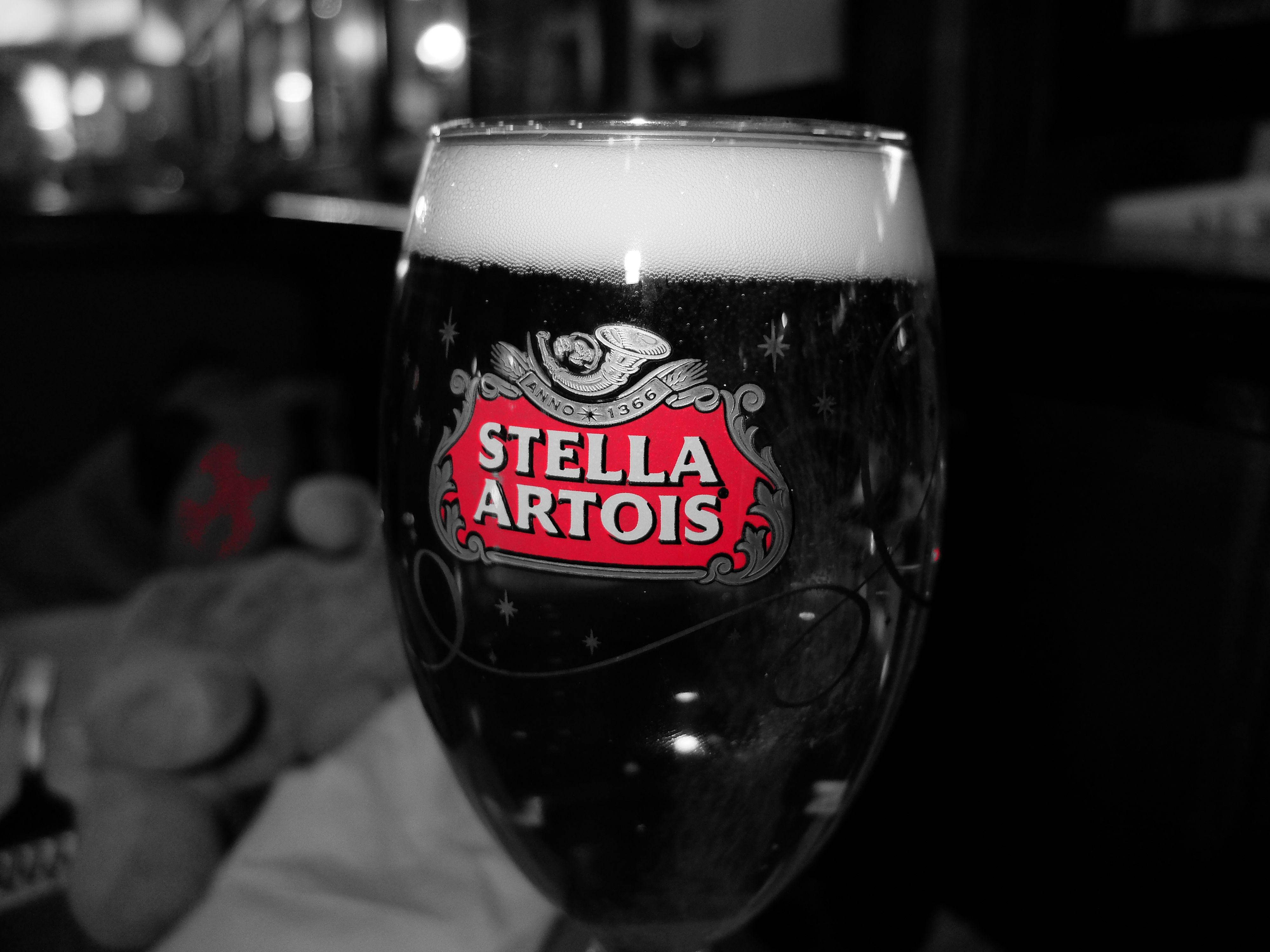 飲料 桌布 壁紙 — 一個黑白特寫的Stella Artois啤酒杯，帶有鮮豔的紅色標誌和泡沫頂部，背景是模糊的酒吧。