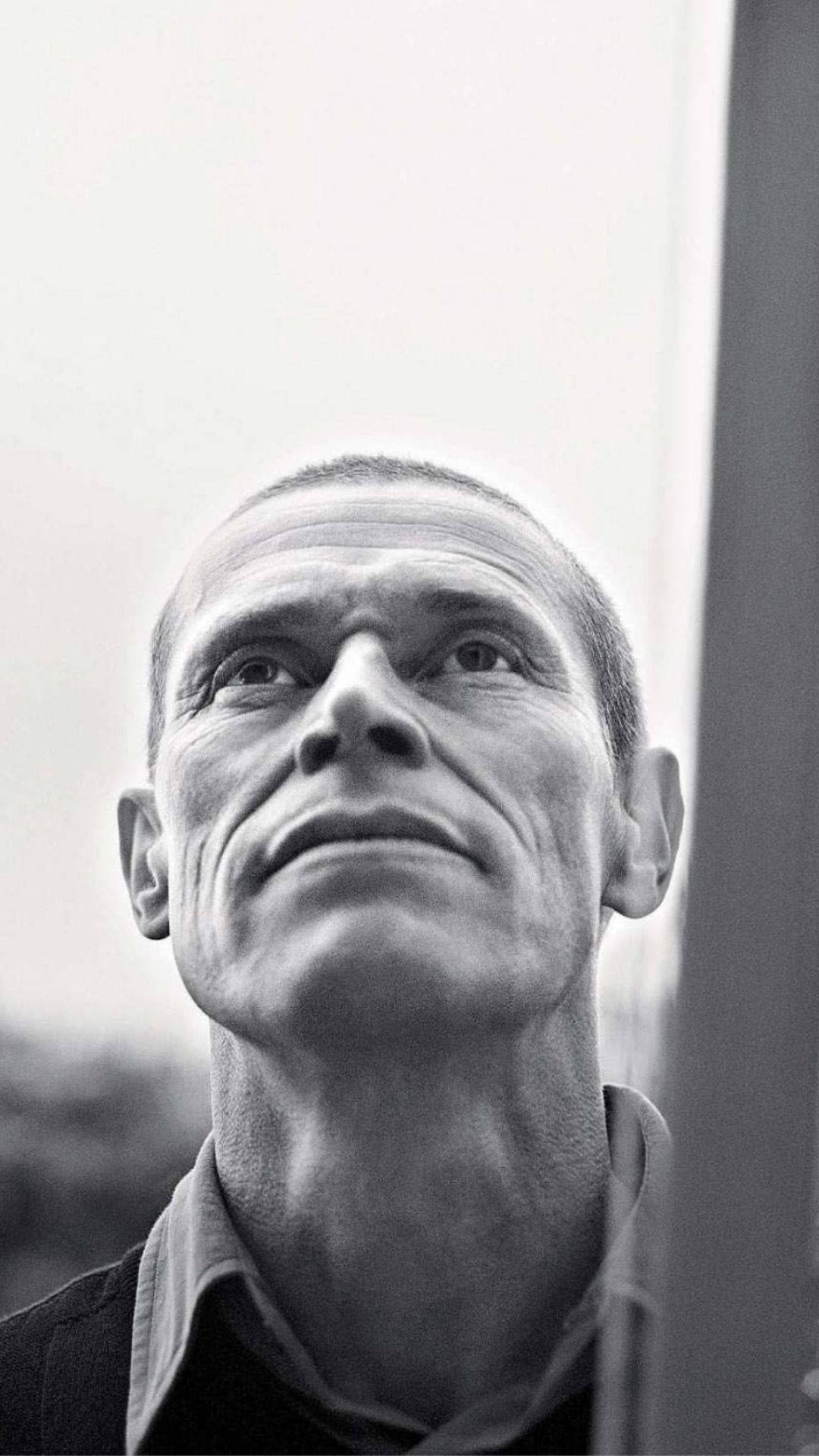 Willem Dafoe 桌布 壁紙 — 一個黑白特寫肖像，一名男子在模糊背景前向上凝視，表情強烈。