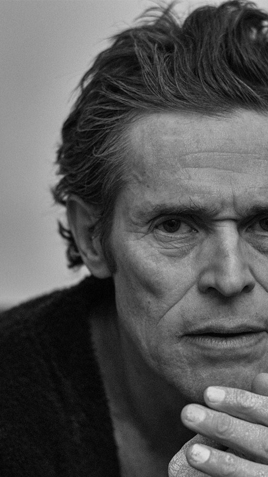 Willem Dafoe 桌布 壁紙 — 一個黑白特寫肖像，拍攝一名頭髮有紋理、皺紋深刻的男子，手放在下巴附近，凝視著鏡頭。