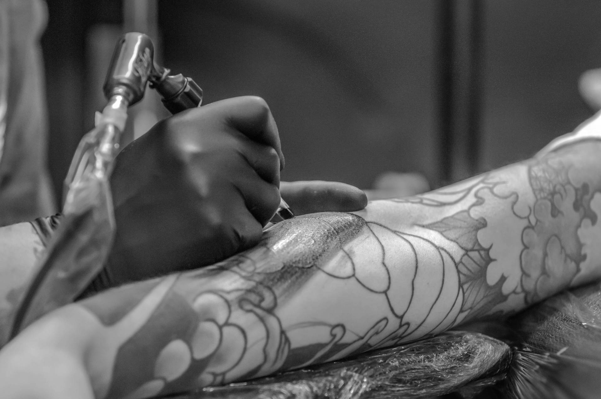 Tattoo 桌布 壁紙 — 一個黑白特寫鏡頭，刺青師傅的手熟練地將墨水注入客戶腿上精緻的花卉圖案。