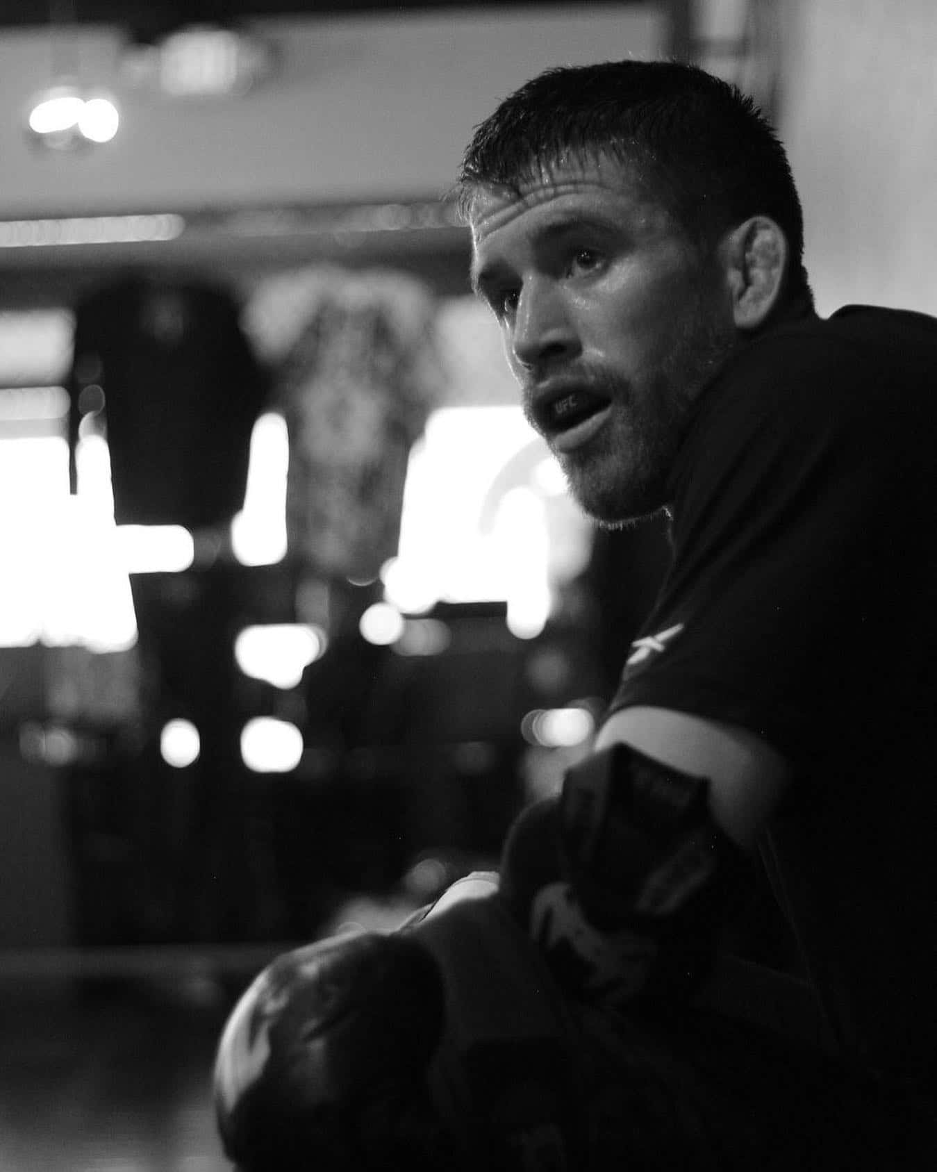 MMA 桌布 壁紙 — 一個黑白特寫鏡頭，拍攝健身房中一名汗流浹背的男子，戴著拳擊手套，專注地凝視鏡頭。