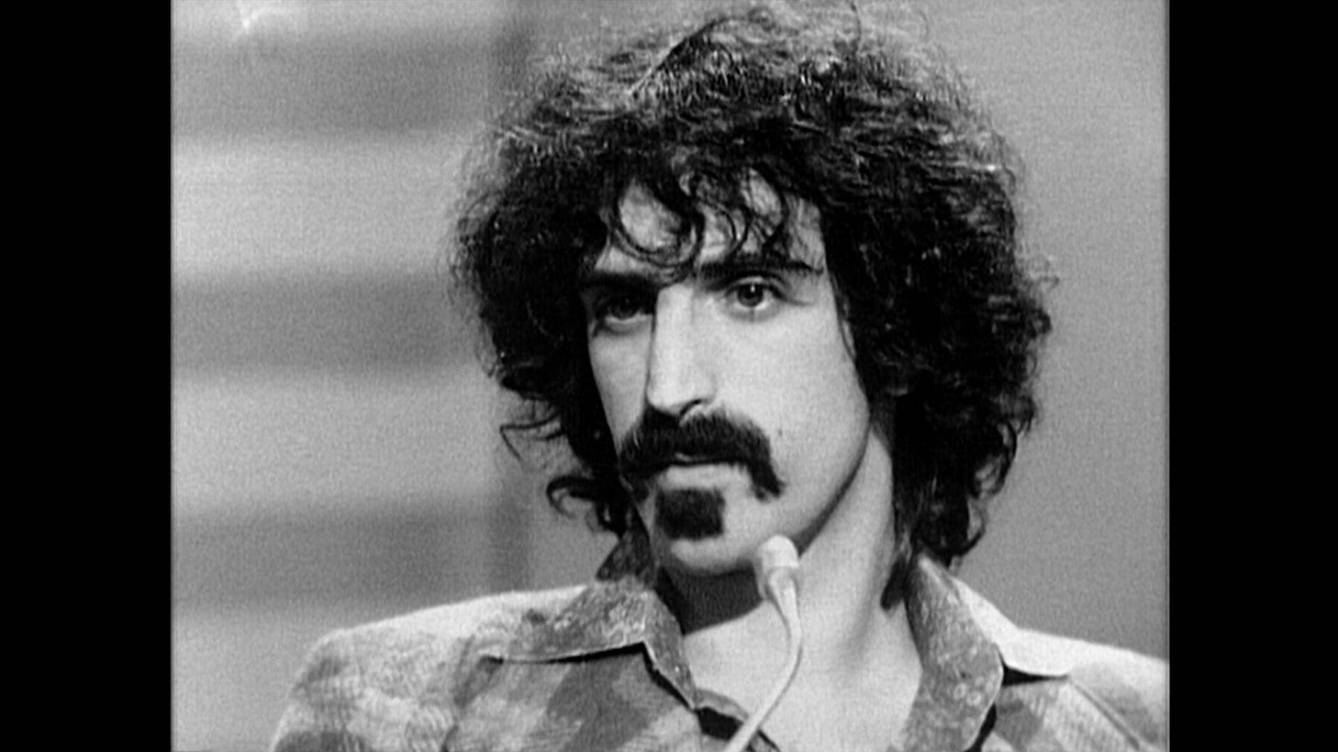 Zappa 桌布 壁紙 — 一個黑白特寫鏡頭，捕捉一名留著捲髮和鬍子的男子對著麥克風說話。
