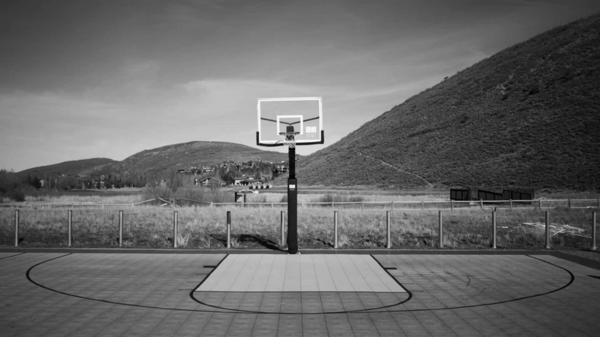 1080p Basketball 桌布 壁紙 — 一個黑白相間的籃球場孤獨地矗立著，背後山巒起伏，天空多雲。