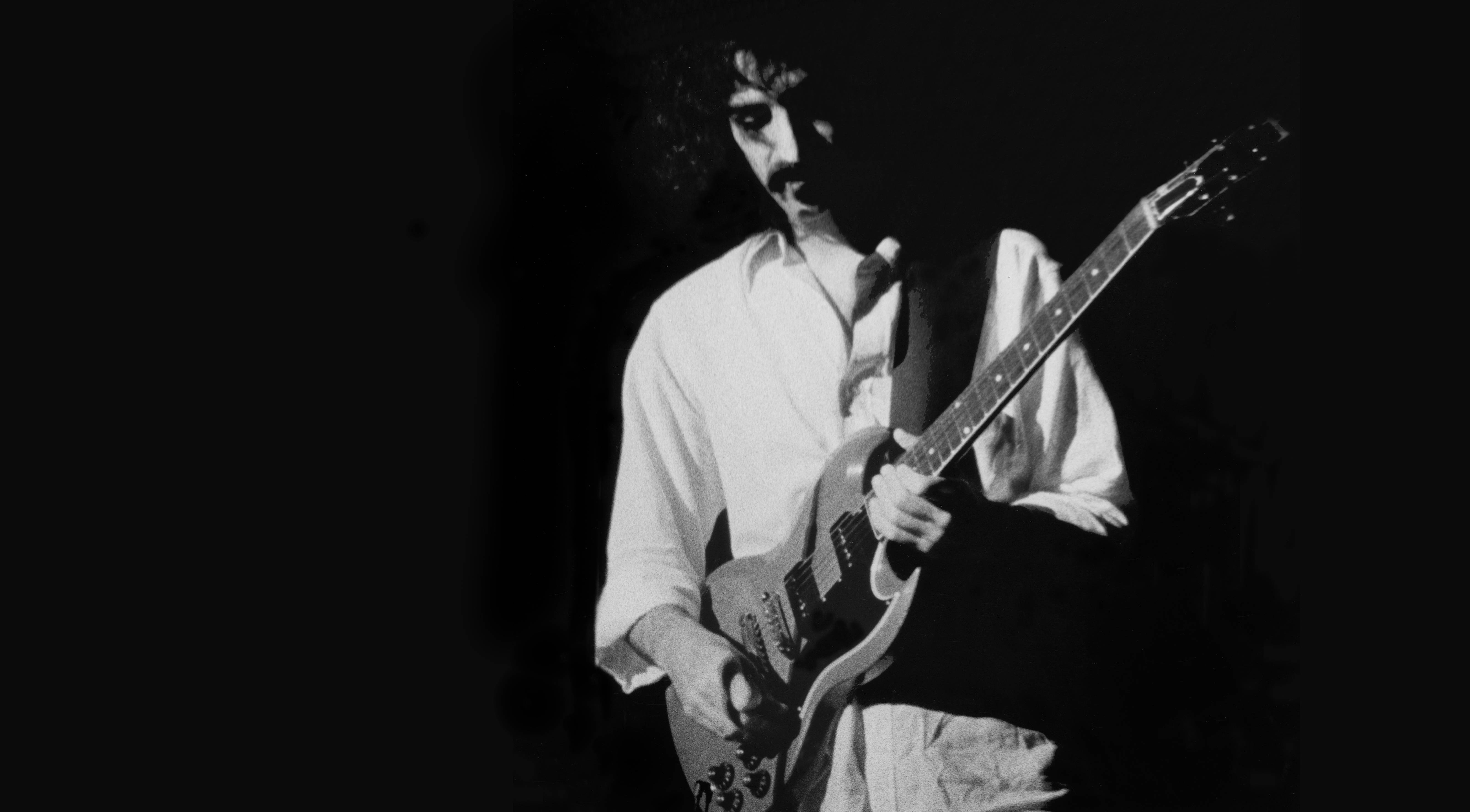 Zappa 桌布 壁紙 — 一個黑白肖像，一個捲髮男子在黑暗背景下彈奏電吉他。