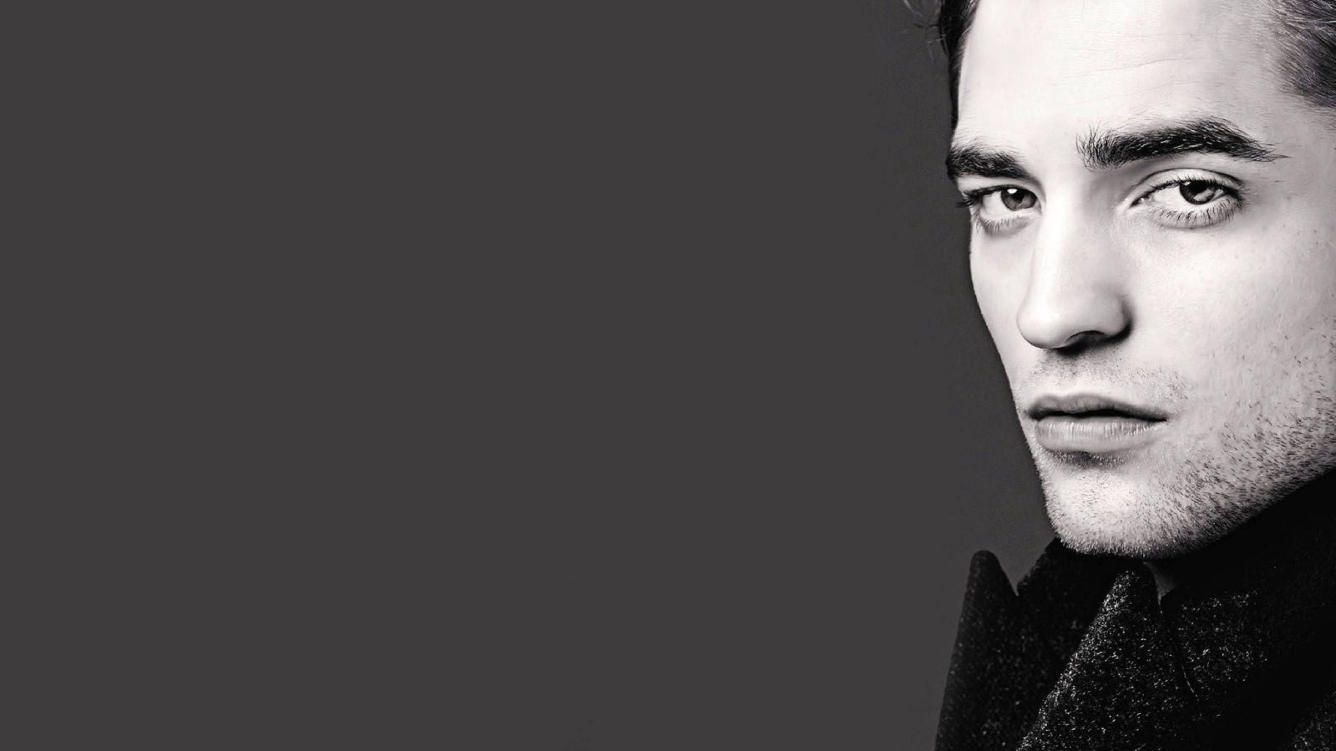 Robert Pattinson 桌布 壁紙 — 一個黑白肖像，一個穿著深色外套的男人在純灰色背景前專注地凝視。