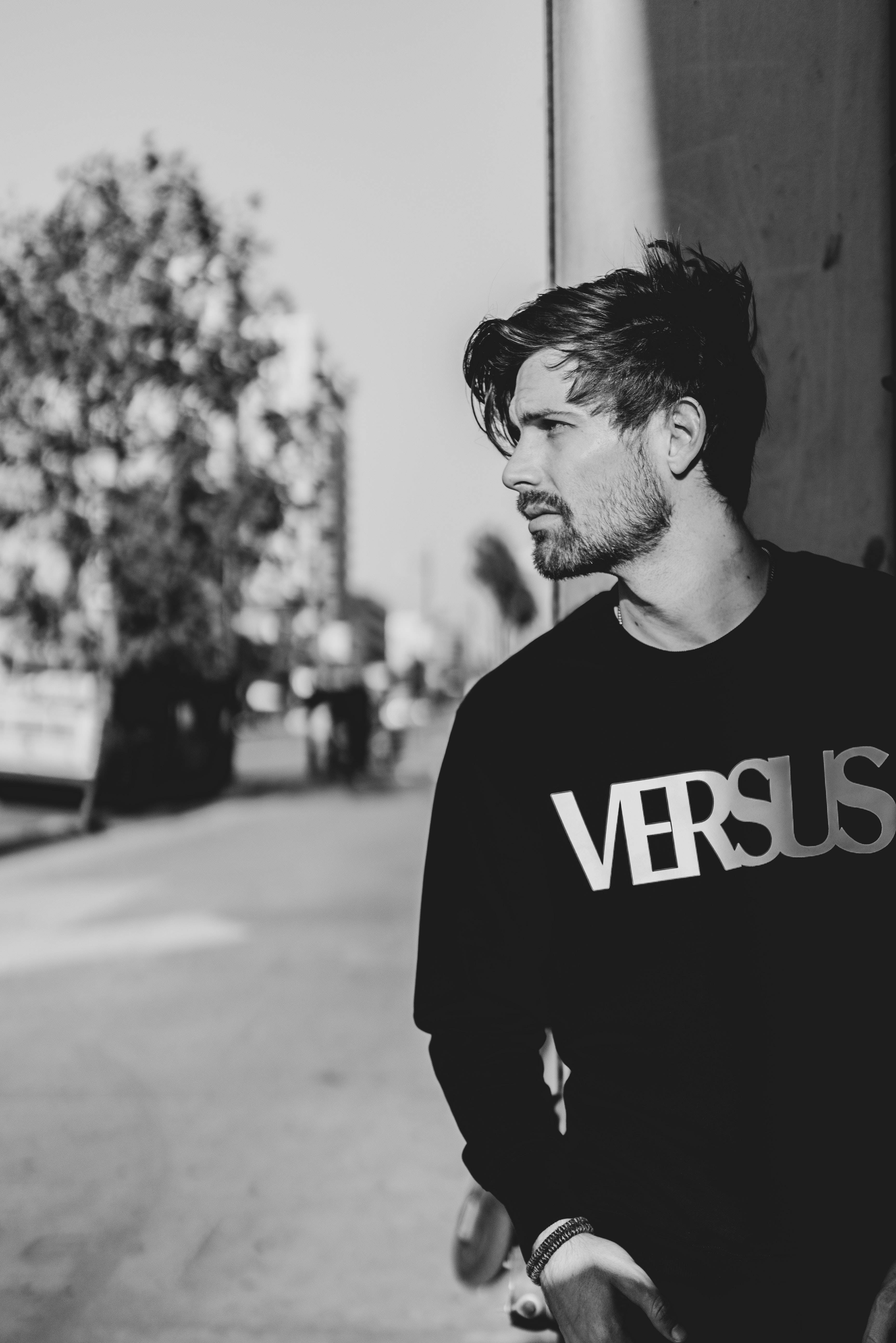 male model 桌布 壁紙 — 一個黑白肖像，一名男子留著時尚髮型和鬍鬚，穿著 'VERSUS' 運動衫，站在陽光照耀的城市街道上。