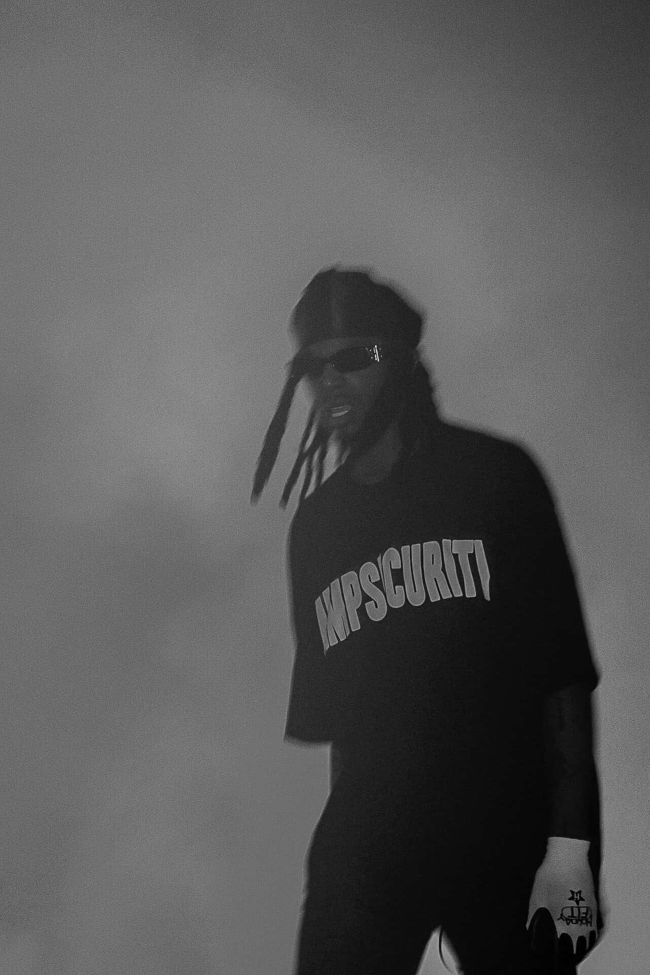 Playboi Carti 桌布 壁紙 — 一個黑白肖像，一名男子留著辮子頭、戴太陽眼鏡，身穿「KMPSCURITI」襯衫和手套，背景煙霧繚繞。