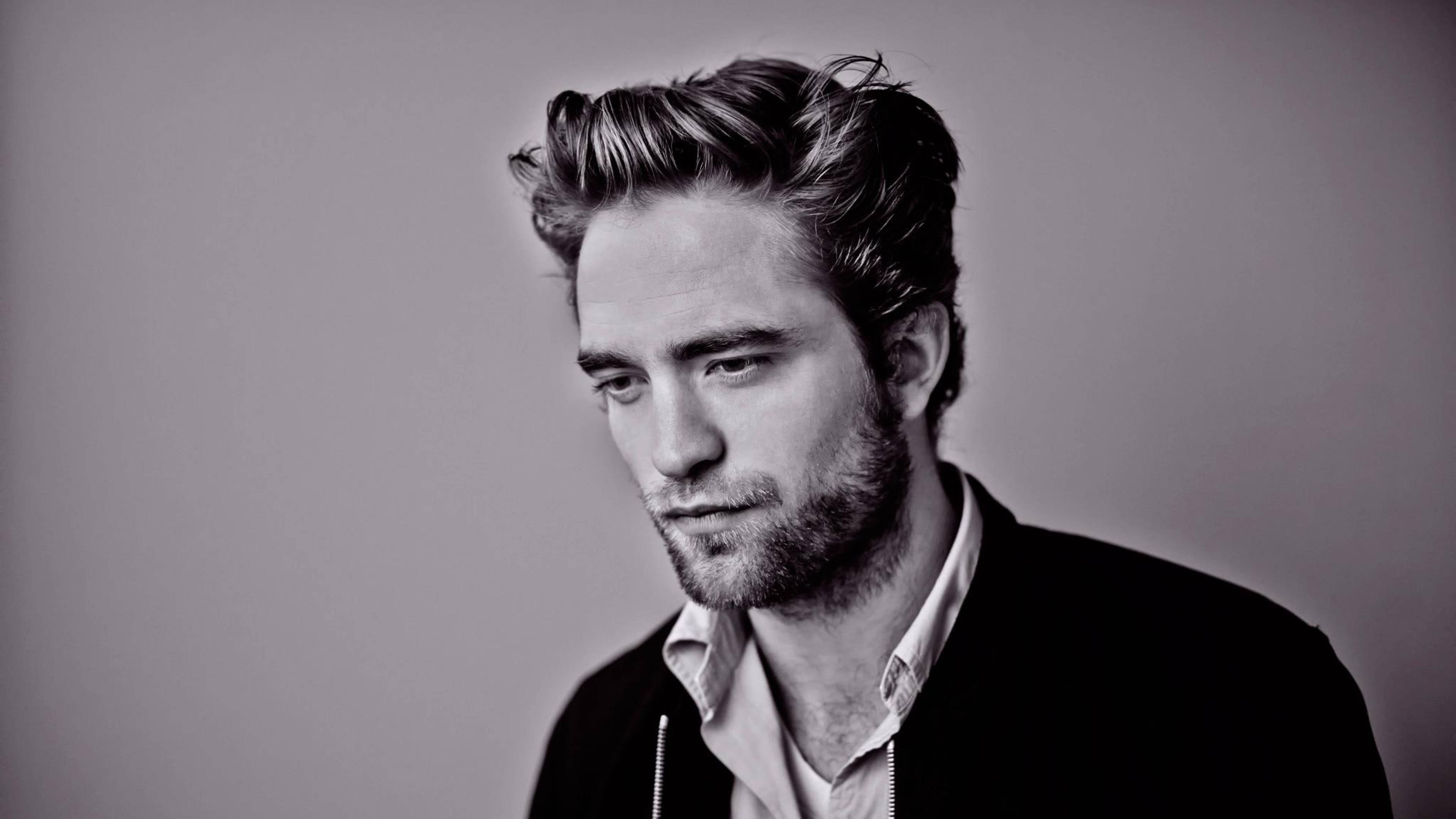 Robert Pattinson 桌布 壁紙 — 一個黑白肖像，一名男子留著造型頭髮和鬍鬚，若有所思地向下看，背景是純灰色。