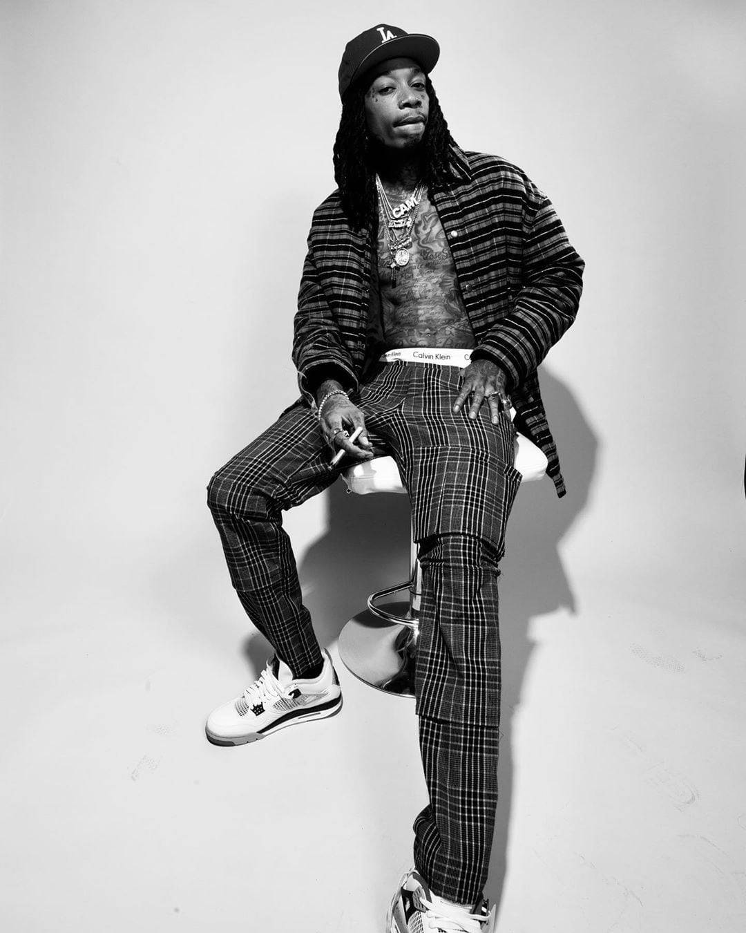 wiz khalifa 桌布 壁紙 — 一個黑白肖像，一名留著辮子的男人坐在凳子上，穿著格子服裝和帽子。
