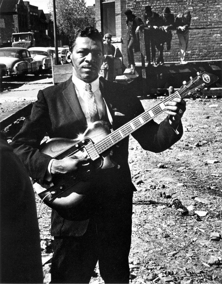 Little Walter 桌布 壁紙 — 一個黑白肖像，一名穿著西裝的男子在骯髒的城市街道上拿著電吉他，背景有復古汽車和旁觀者。