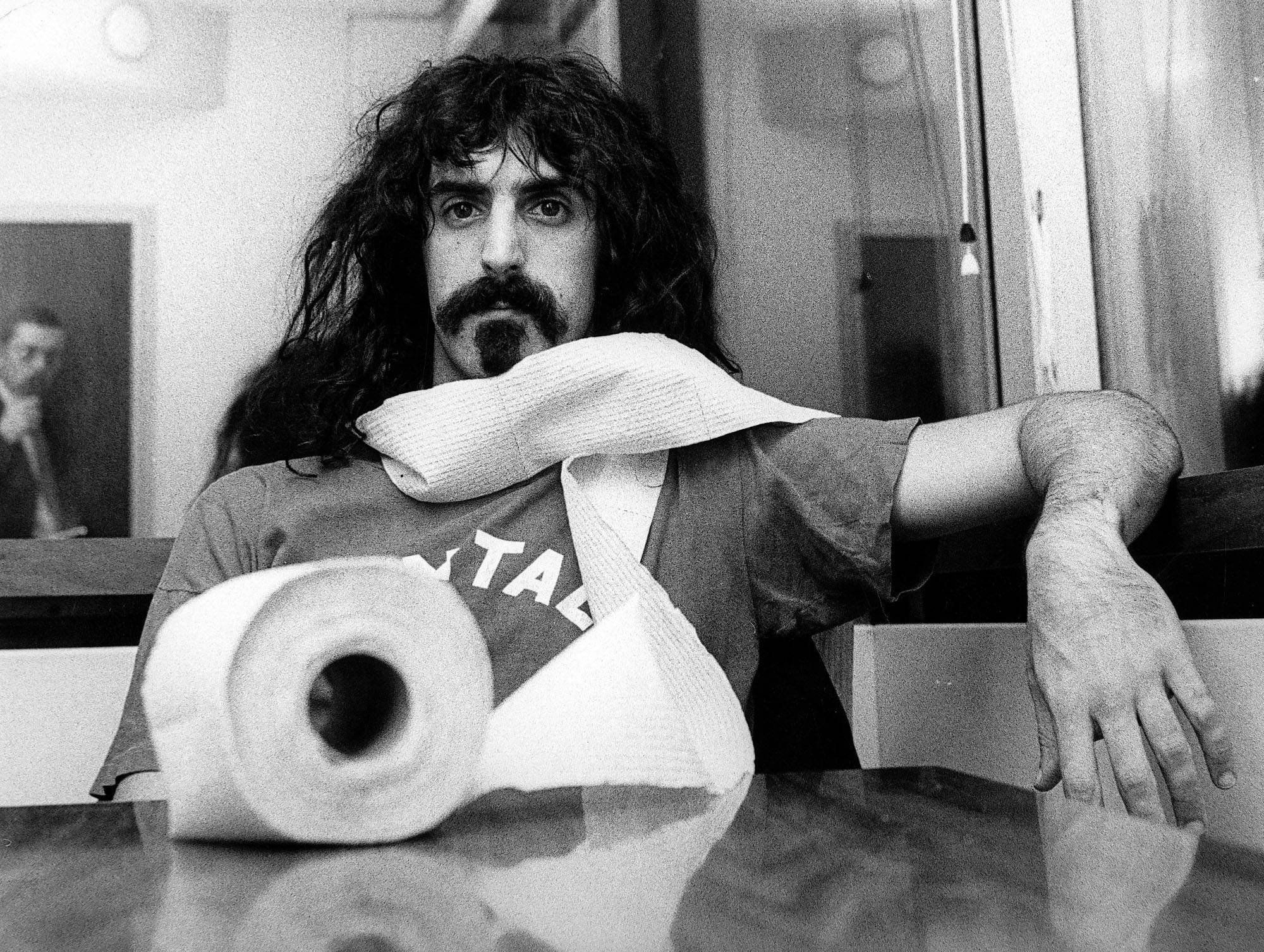 Zappa 桌布 壁紙 — 一個黑白肖像，一名長髮鬍鬚男子倚在放有衛生紙卷和展開紙張的桌子上。