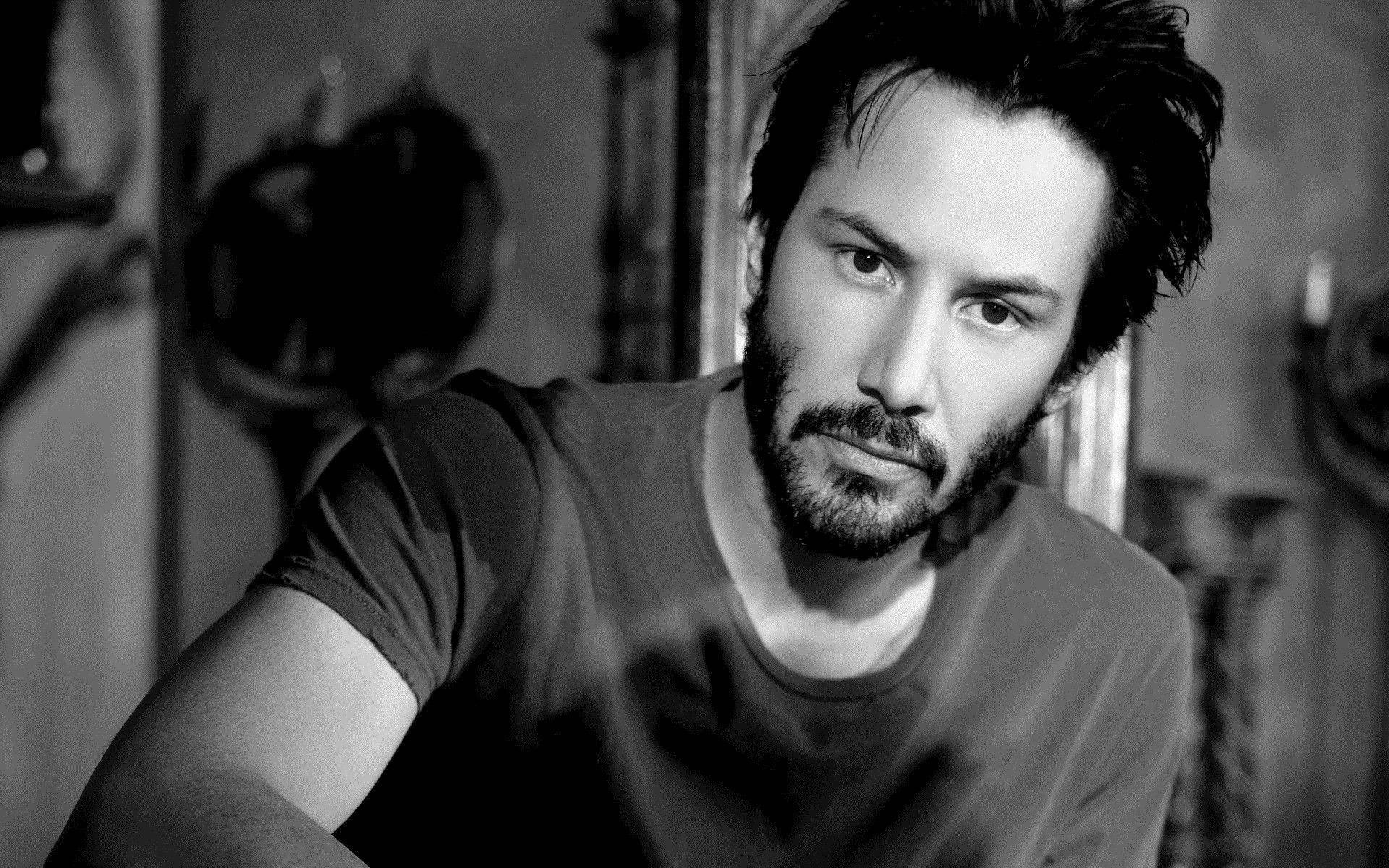 Keanu 桌布 壁紙 — 一個黑白色調的肖像，拍攝一位深色頭髮和鬍鬚的男人，在昏暗的房間裡專注地看著相機。