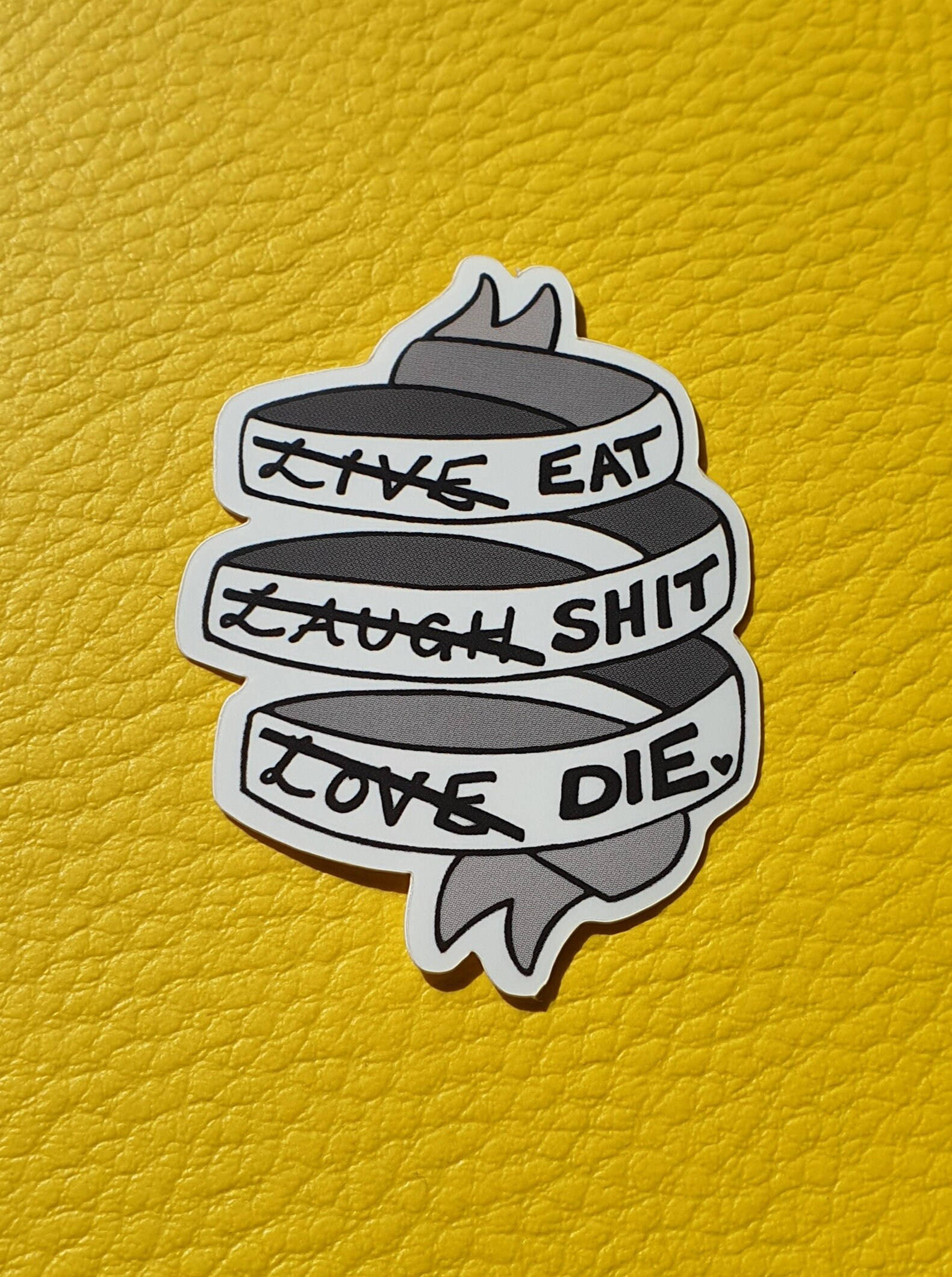 藝術 桌布 壁紙 — 一個黑白貼紙，上面有被劃掉的短語 'live eat'、'laugh shit'、'love die'，位於黃色紋理表面上。